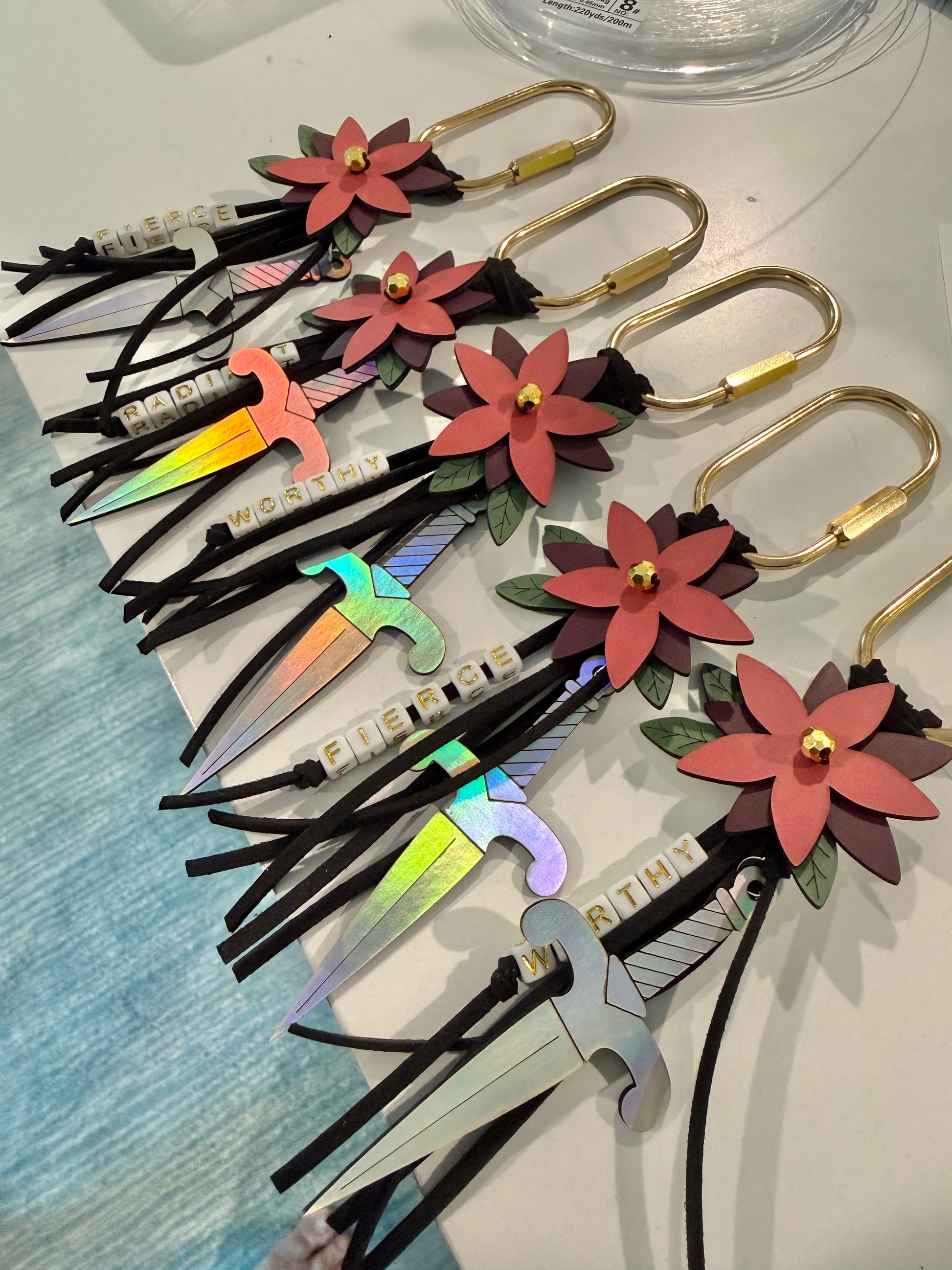 Bloom & Dagger romantasy bag charms by zoo&roo with layered burgundy vegan leather flower, iridescent dagger charm, green leatherette leaves, and words WORTHY, FIERCE,  and RADIANT spelled out in beads on gold brass carabiner.