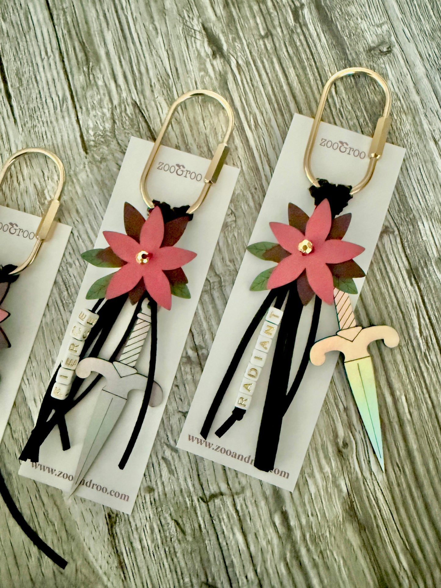 Bloom & Dagger romantasy bag charms by zoo&roo with layered burgundy vegan leather flower, iridescent dagger charm, green leatherette leaves, and words FIERCE and RADIANT spelled out in beads on gold brass carabiner