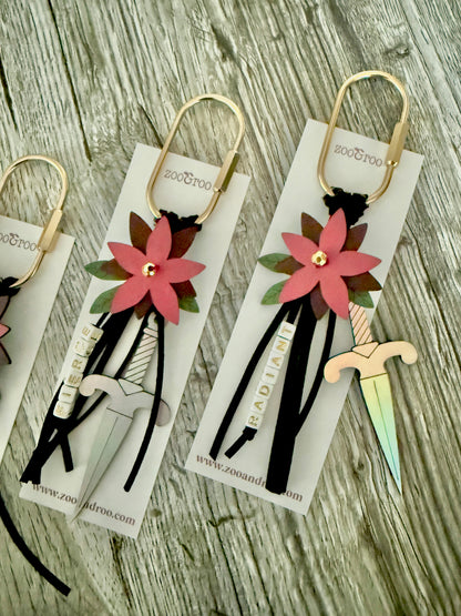 Bloom & Dagger romantasy bag charms by zoo&roo with layered burgundy vegan leather flower, iridescent dagger charm, green leatherette leaves, and words FIERCE and RADIANT spelled out in beads on gold brass carabiner