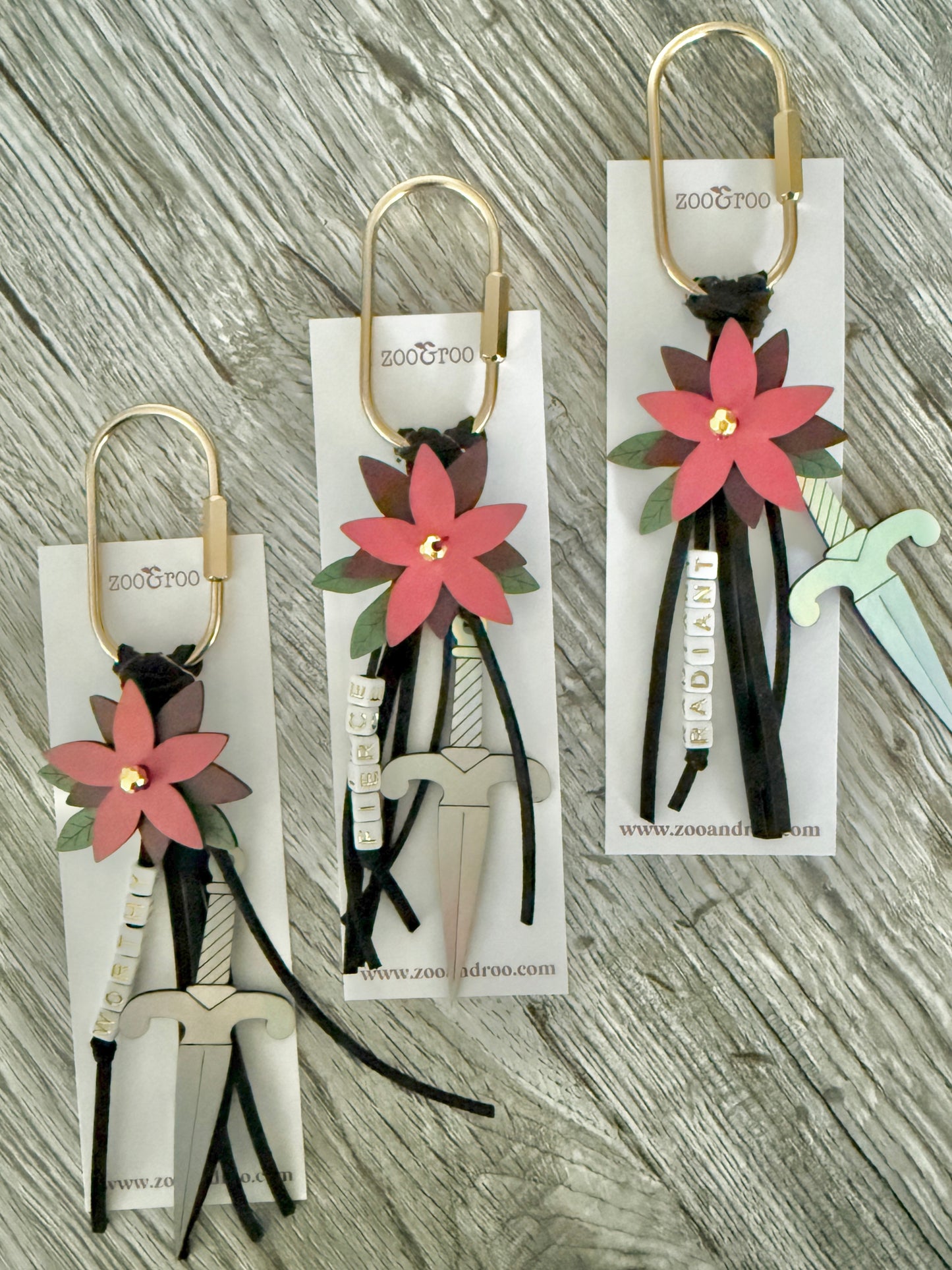 Bloom & Dagger romantasy bag charms by zoo&roo with layered burgundy vegan leather flower, iridescent dagger charm, green leatherette leaves, and words WORTHY, FIERCE,  and RADIANT spelled out in beads on gold brass carabiner