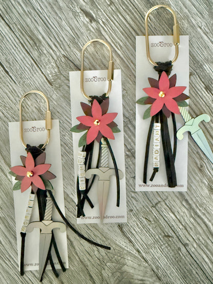 Bloom & Dagger romantasy bag charms by zoo&roo with layered burgundy vegan leather flower, iridescent dagger charm, green leatherette leaves, and words WORTHY, FIERCE,  and RADIANT spelled out in beads on gold brass carabiner