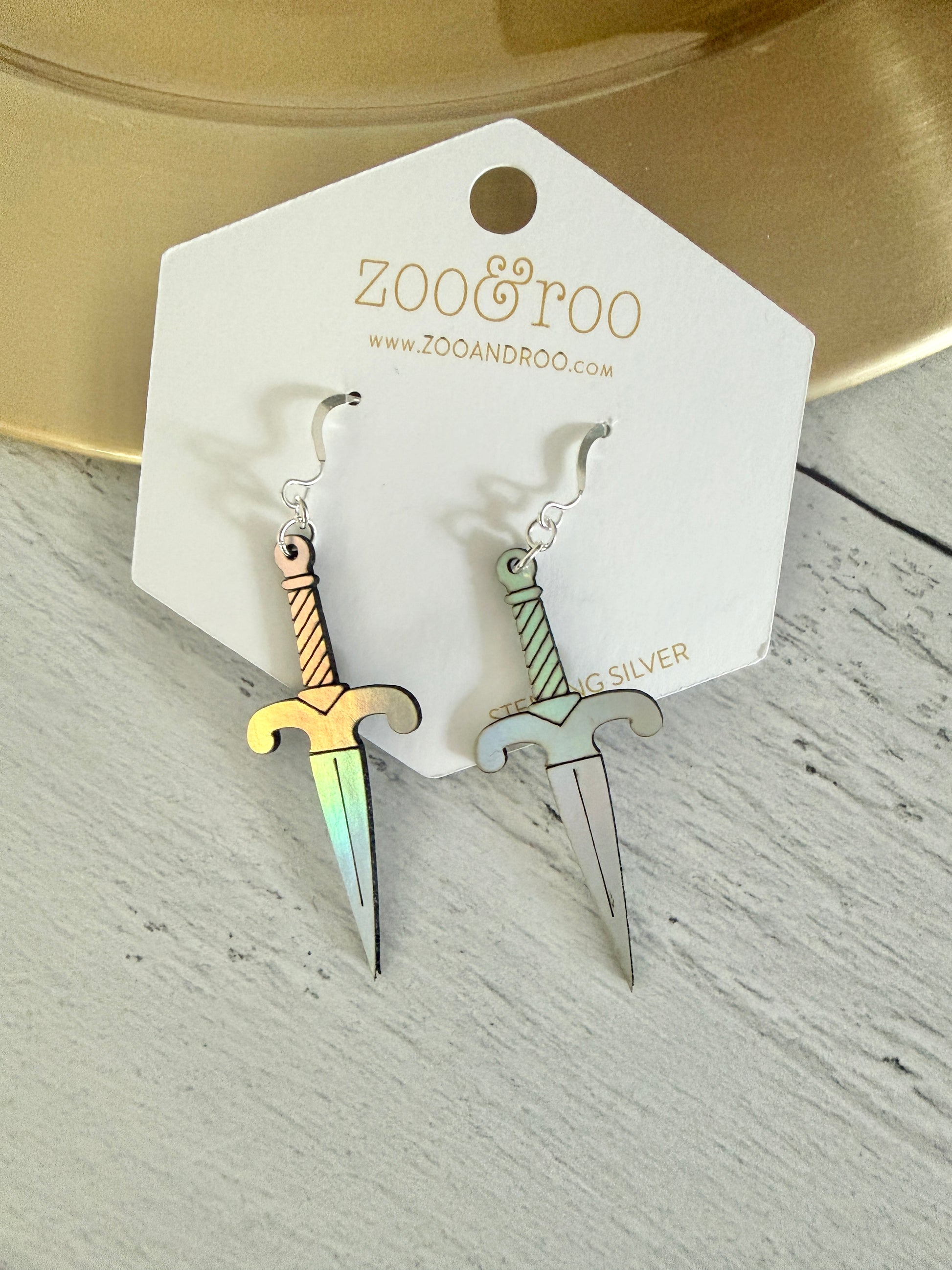 A pair of iridescent vegan leather dagger earrings, displayed on a white zoo&roo brand card against a light gray wood background.
