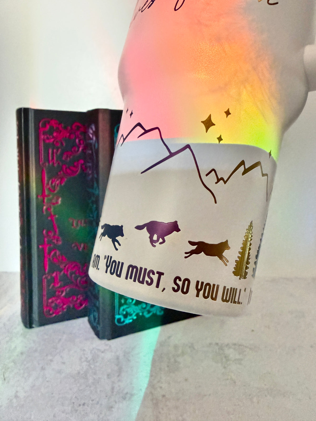 Closeup of a white 40 oz Dire Bound inspired tumbler with rainbow-engraved wolves running around the base of the tumbler with mountains, pine trees and stars and the quote 'you must, so you will.'