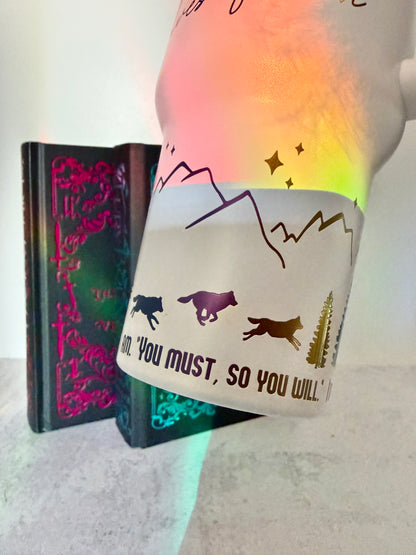Closeup of a white 40 oz Dire Bound inspired tumbler with rainbow-engraved wolves running around the base of the tumbler with mountains, pine trees and stars and the quote 'you must, so you will.'
