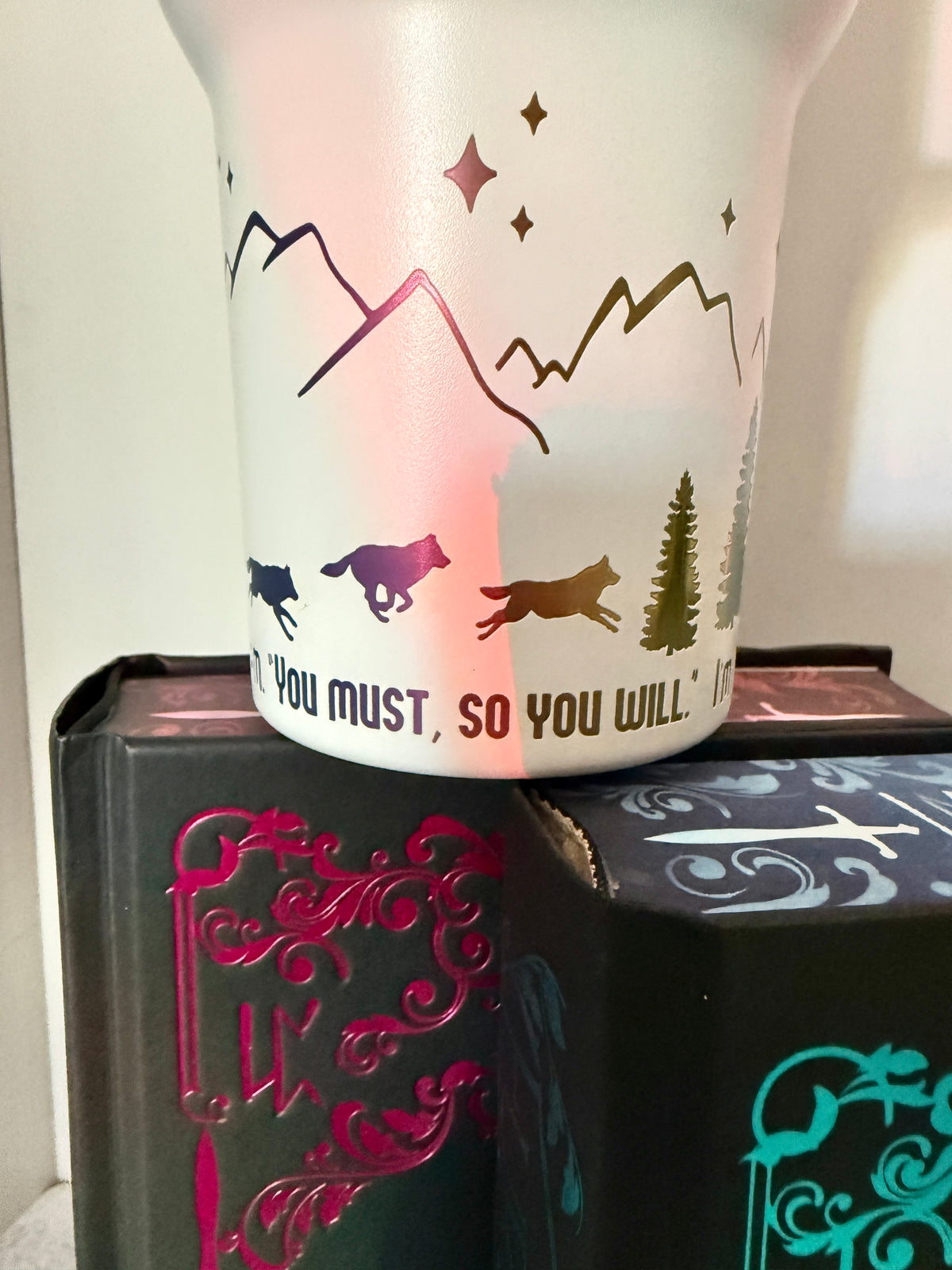 Closeup of a white 40 oz Dire Bound inspired tumbler with rainbow-engraved wolves running around the base of the tumbler with mountains, pine trees and stars and the quote 'you must, so you will.'
