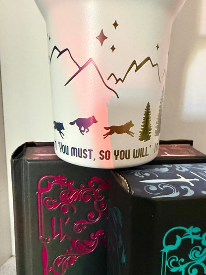 Closeup of a white 40 oz Dire Bound inspired tumbler with rainbow-engraved wolves running around the base of the tumbler with mountains, pine trees and stars and the quote 'you must, so you will.'