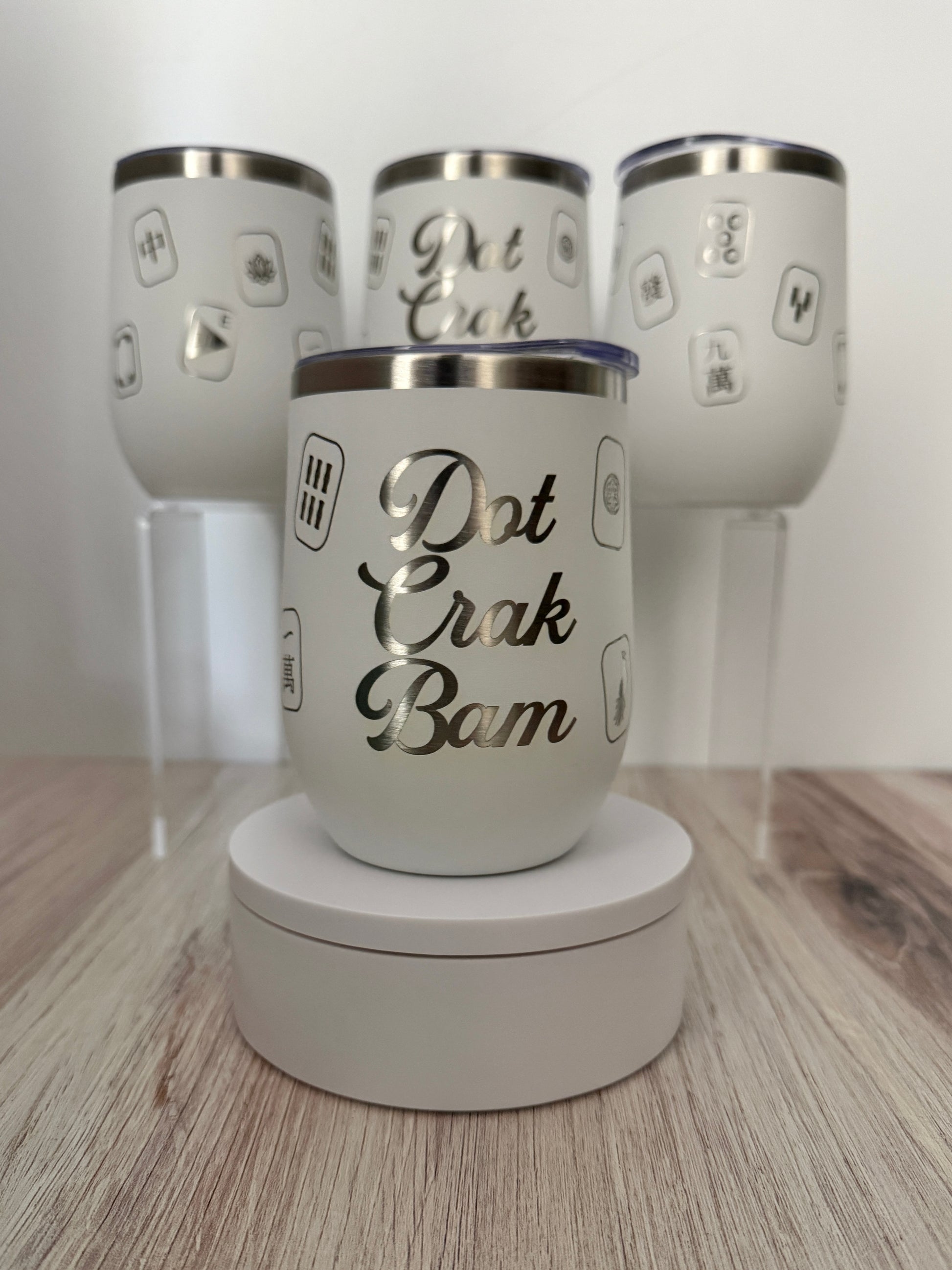 A white insulated mahjong wine tumbler with a silver lid sits on a round white display base in front of three matching tumblers on clear stands. The front tumbler has the phrase “Dot Crak Bam” in large engraved script, with smaller mahjong tiles engraved around the sides.