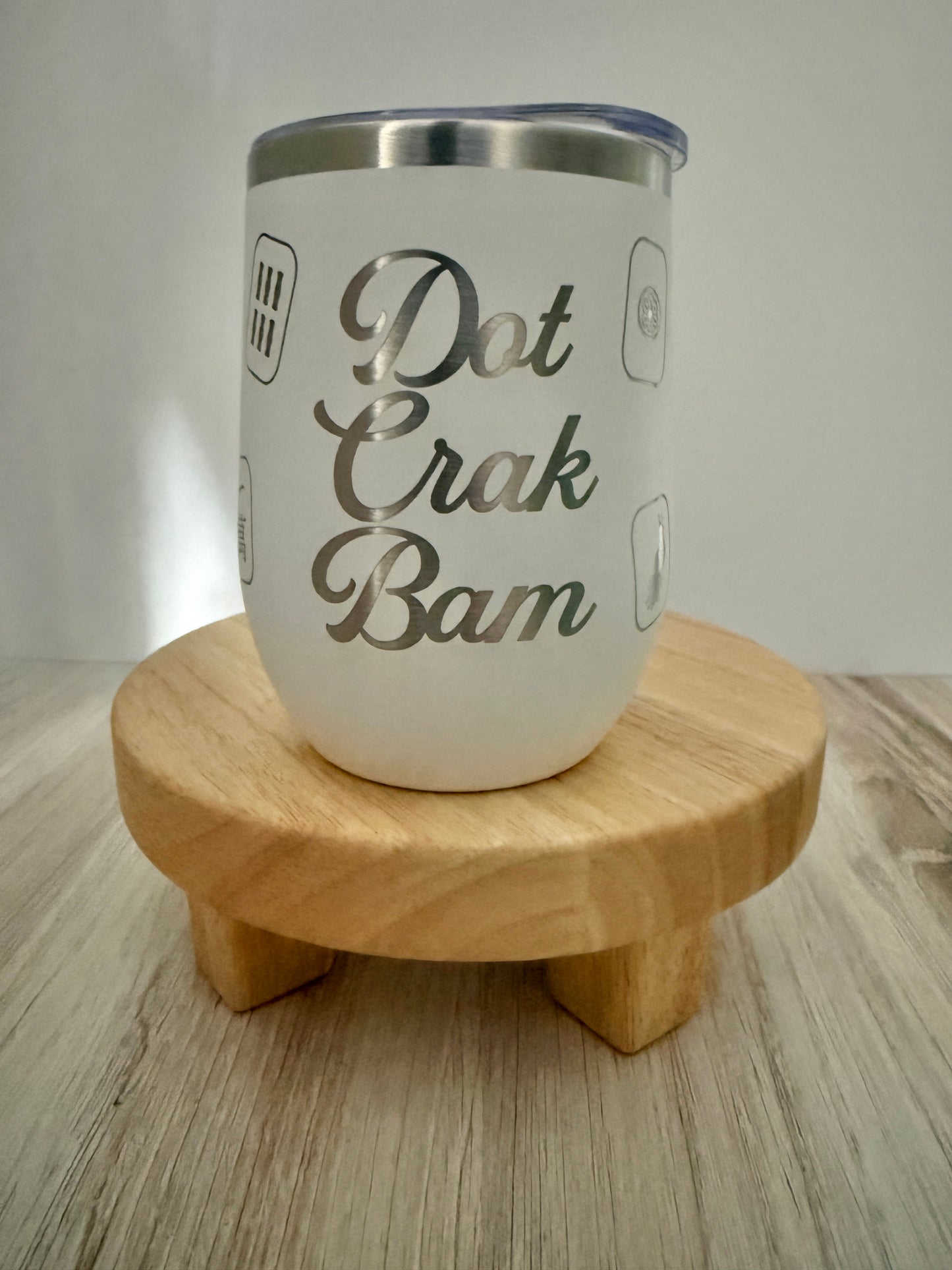 A white insulated mahjong wine tumbler with a stainless steel rim and a clear lid, displayed on a small round wooden stand. The tumbler features the phrase 'Dot Crak Bam', with small mahjong tiles engraved on the sides.