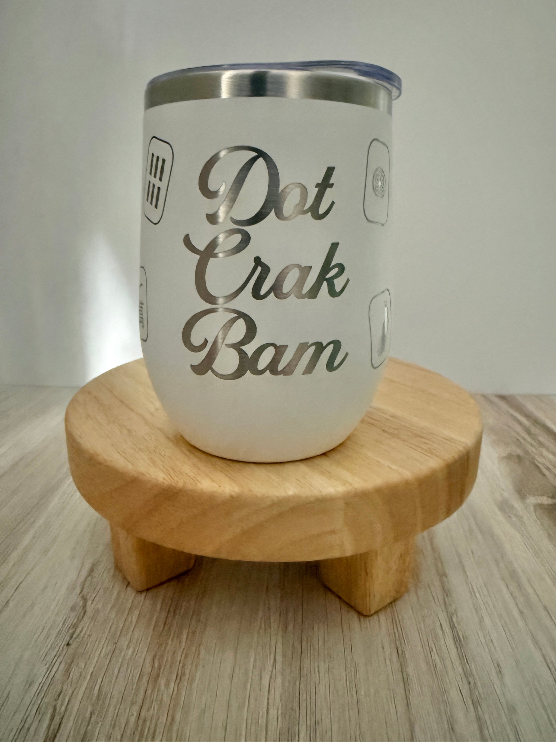 A white insulated mahjong wine tumbler with a stainless steel rim and a clear lid, displayed on a small round wooden stand. The tumbler features the phrase 'Dot Crak Bam', with small mahjong tiles engraved on the sides.