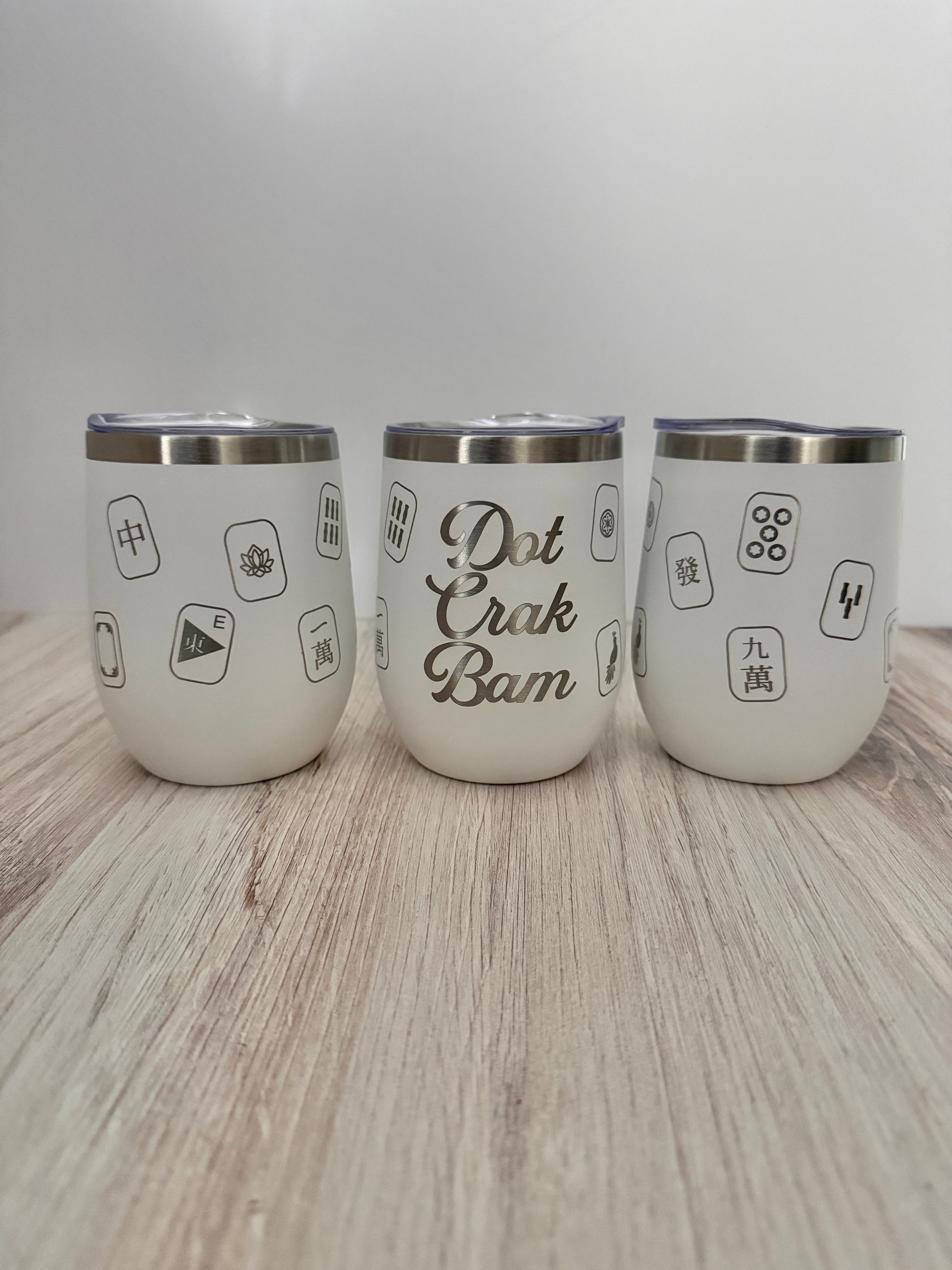 Three white stainless steel insulated mahjong wine tumblers with lids arranged on a light wood surface against a white background. The center tumbler reads 'Dot Crak Bam' engraved with mahjong tiles wrapped around the sides.