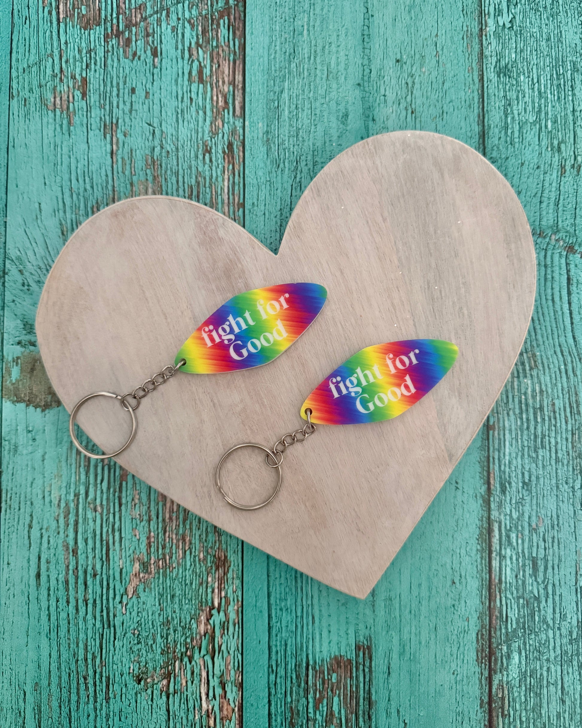 Two rainbow-colored keychains with 'Fight for Good' text on a heart-shaped wooden stand against a teal wooden background.