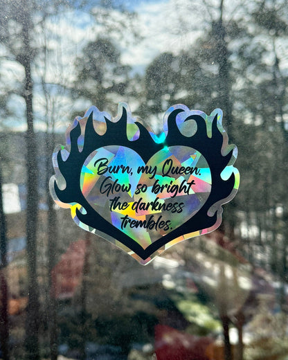 Holographic heart-shaped suncatcher decal surrounded by flames with text "burn, my queen" on a window with a blurred outdoor background