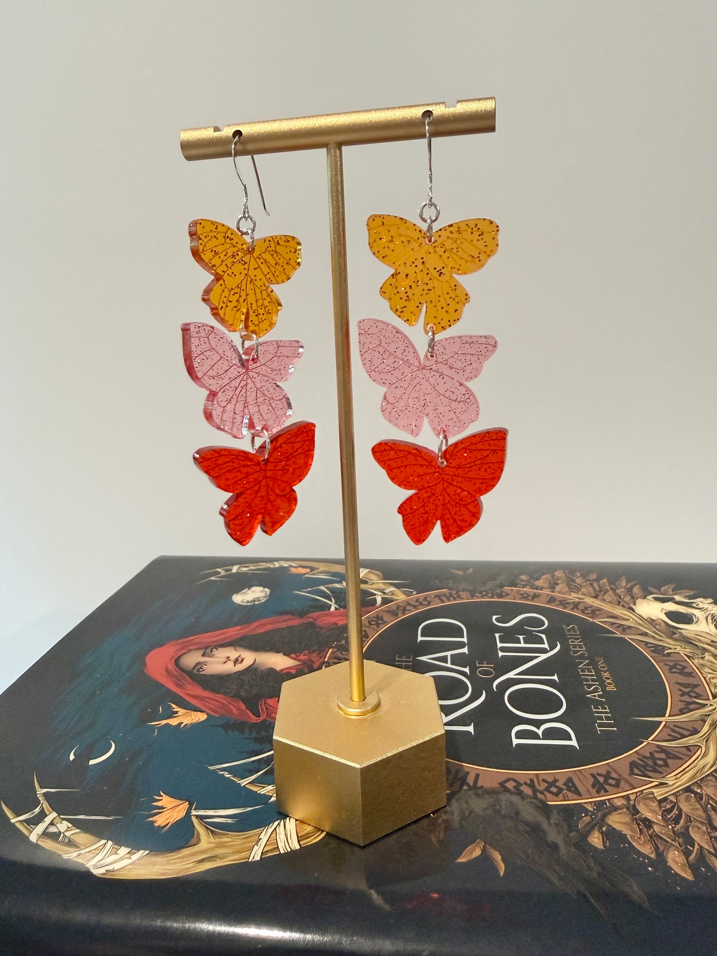 Butterfly-shaped earrings on a gold stand with a book titled 'The Road of Bones' by Demi Winters in the background.