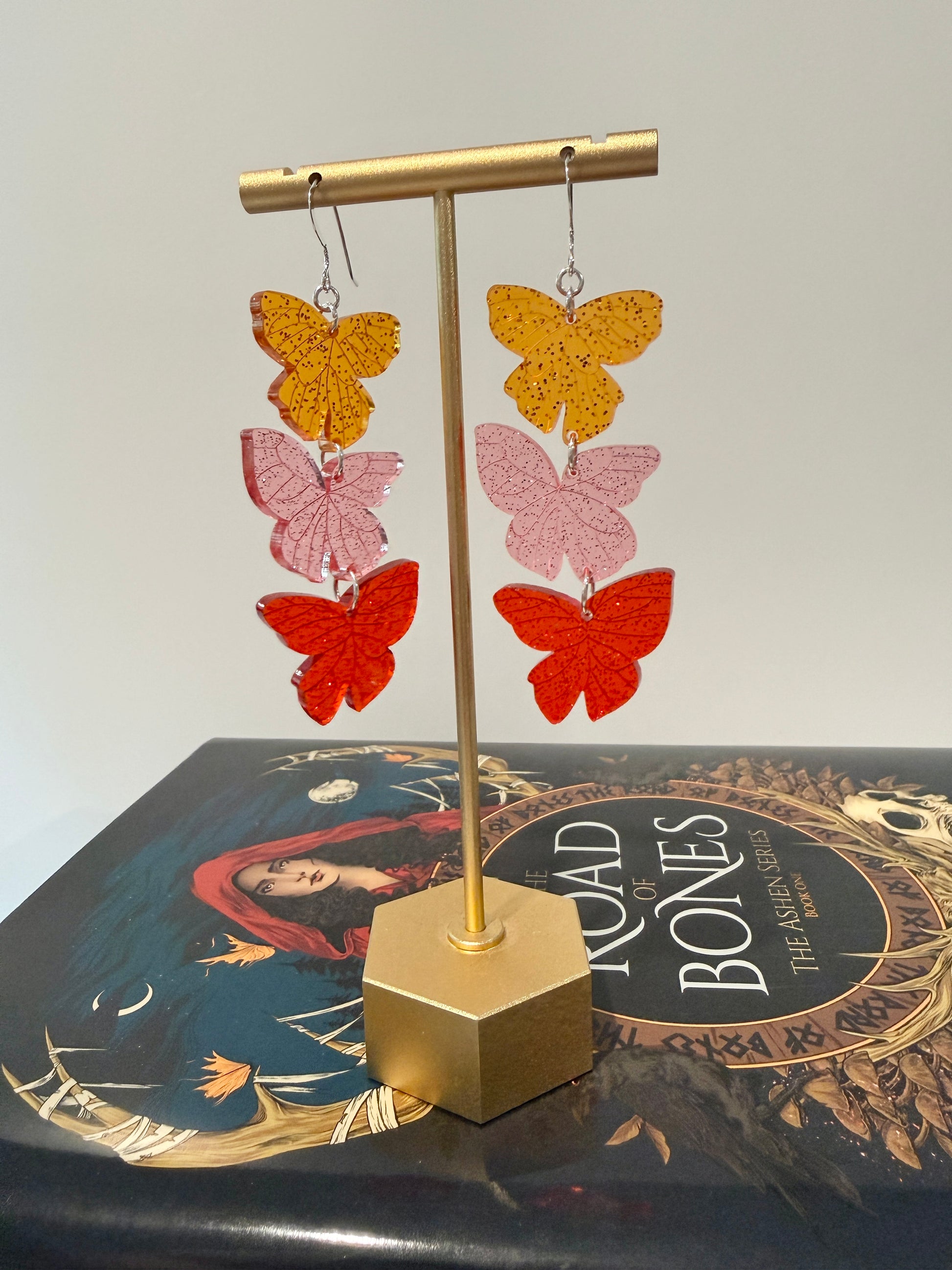 Butterfly-shaped earrings on a gold stand with a book titled 'The Road of Bones' by Demi Winters in the background.