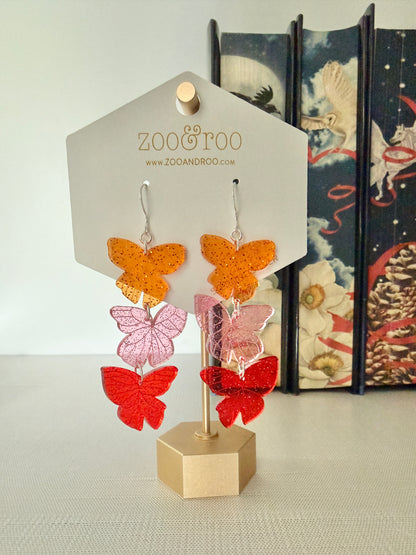 Flíta butterfly earrings inspired by Road of Bones by Demi Winters on a display card with zoo&roo branding, against a decorative book background.