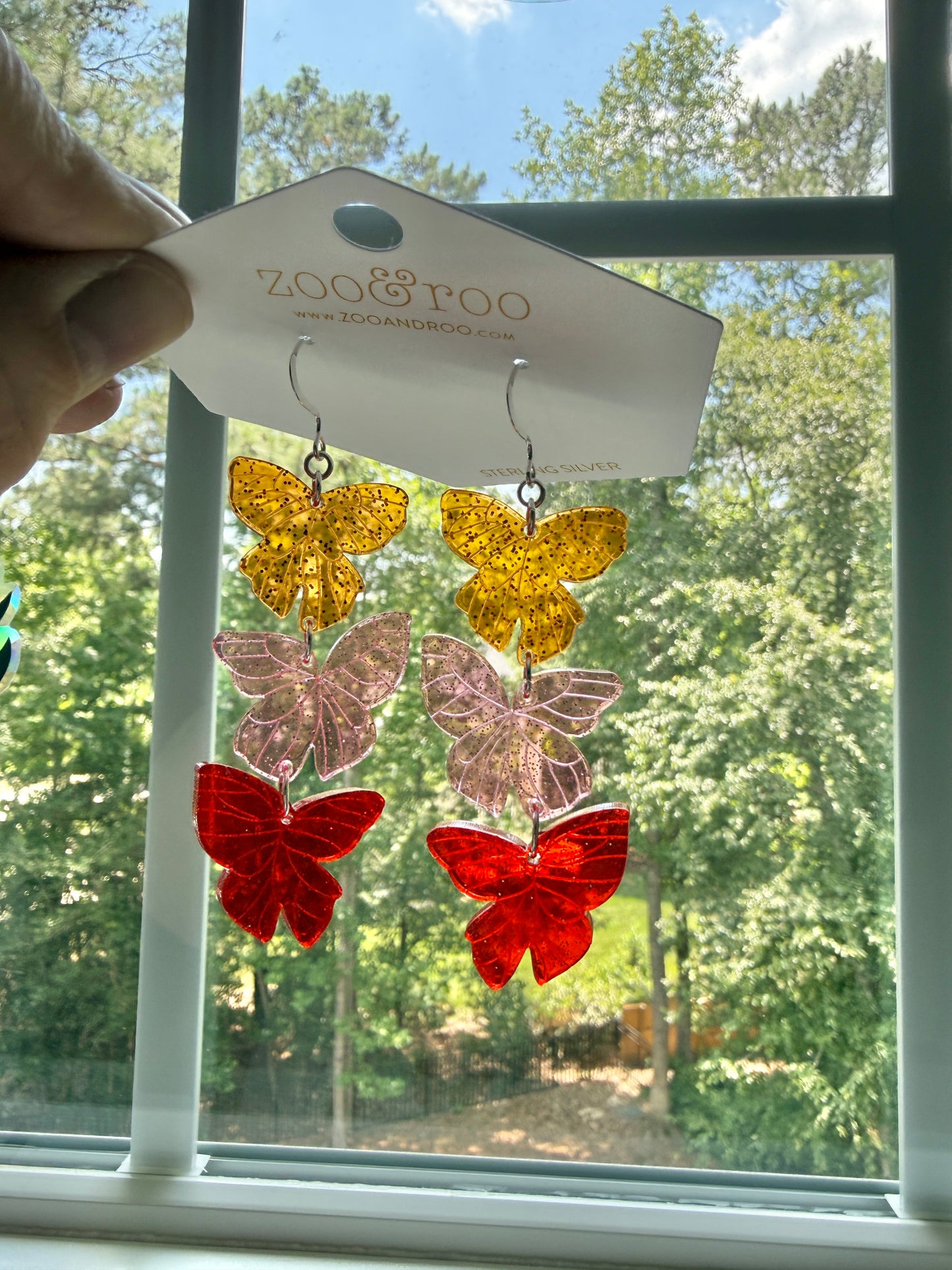 Flíta butterfly earrings inspired by Road of Bones by Demi Winters on a display card with zoo&roo branding, against a window with trees in background.