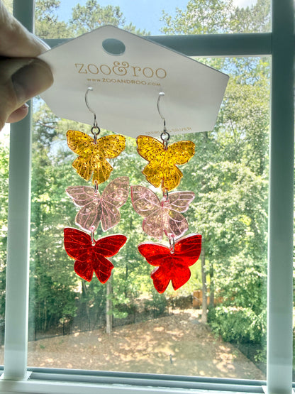 Flíta butterfly earrings inspired by Road of Bones by Demi Winters on a display card with zoo&roo branding, against a window with trees in background.