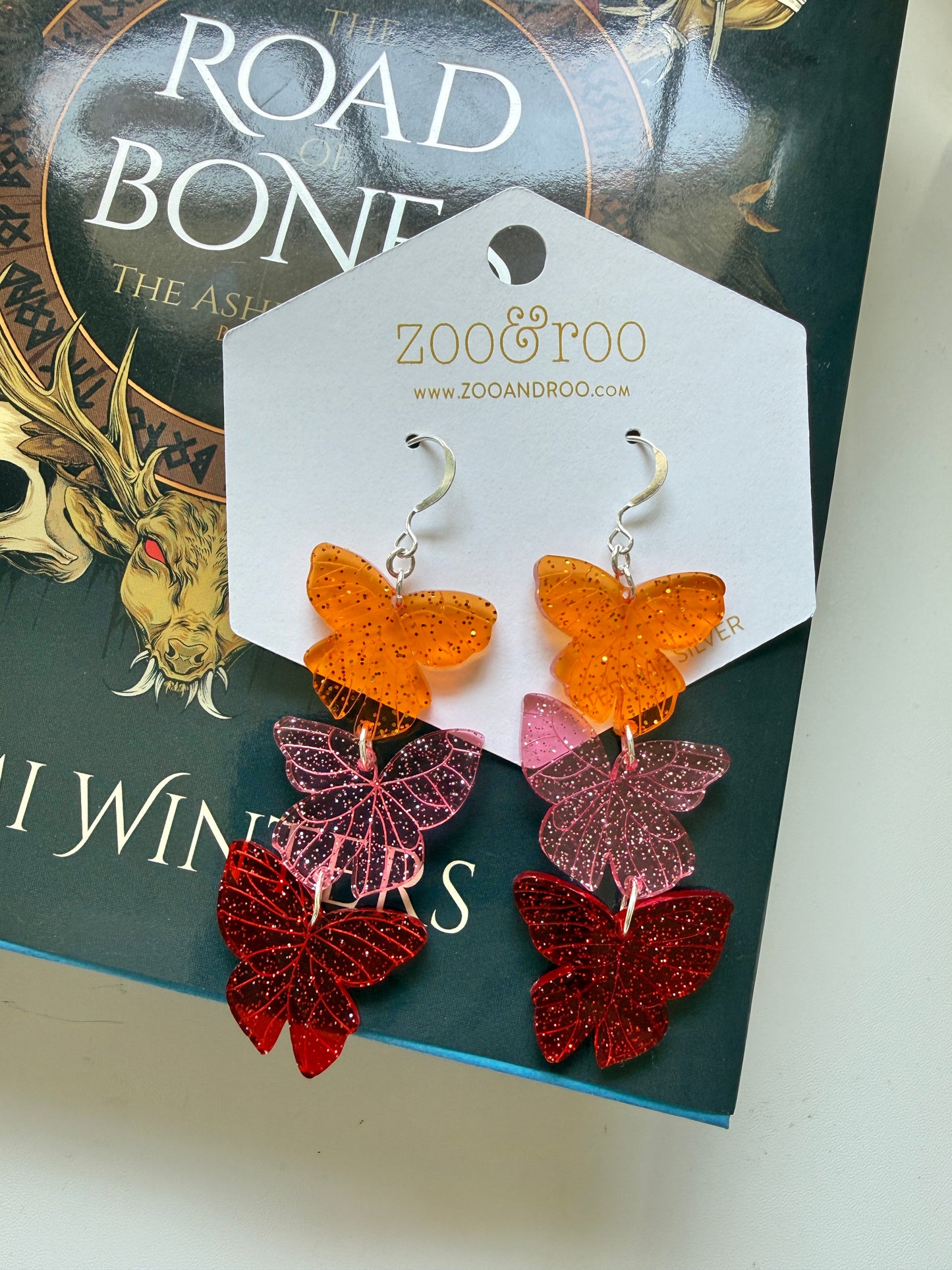 Flíta butterfly earrings inspired by the Ashen series by Demi Winters on a display card with zoo&roo branding, laying on a Road of Bones book.