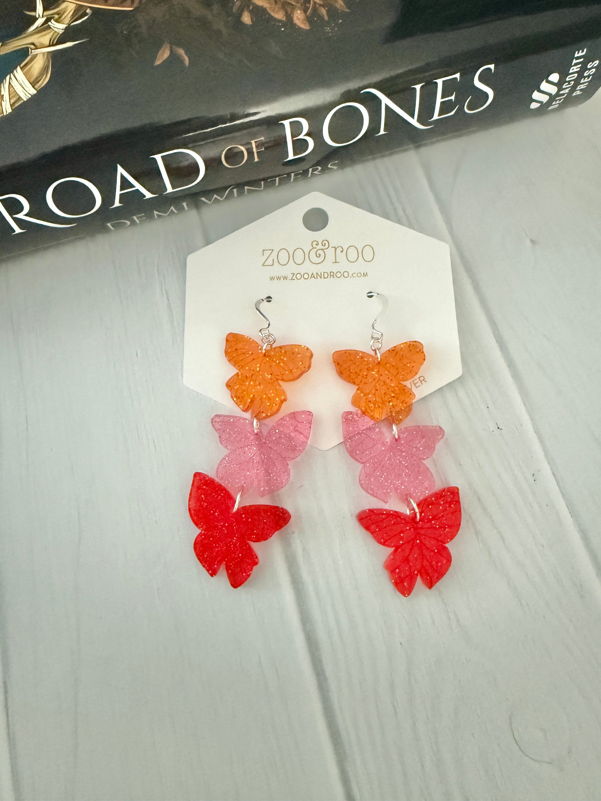 Flíta butterfly earrings inspired by the Ashen series by Demi Winters on a display card with zoo&roo branding, against a Road of Bones book.