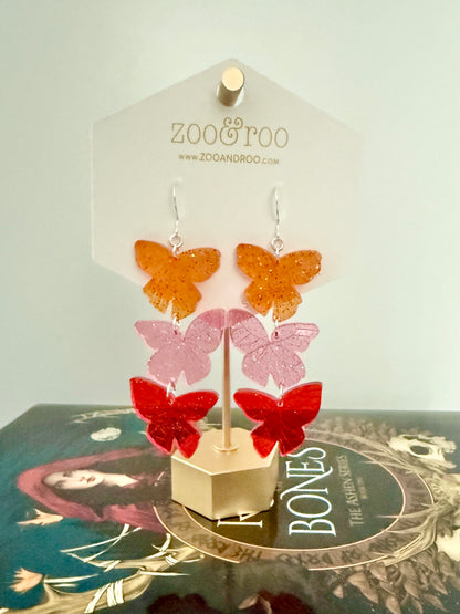 Flíta butterfly earrings inspired by the Ashen series by Demi Winters on a display card with zoo&roo branding, hanging on top of a Road of Bones book.