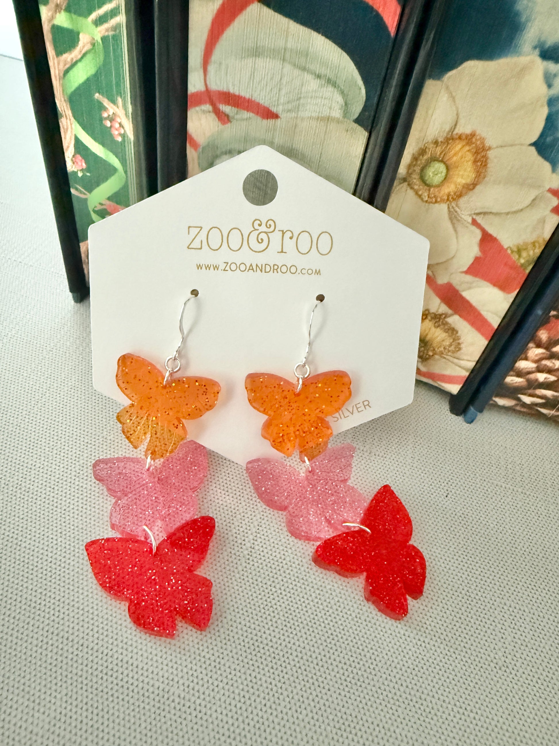 Butterfly-shaped earrings on a ZOO&ROO branded card with a decorative book background, inspired by the Road of Bones by Demi Winters.