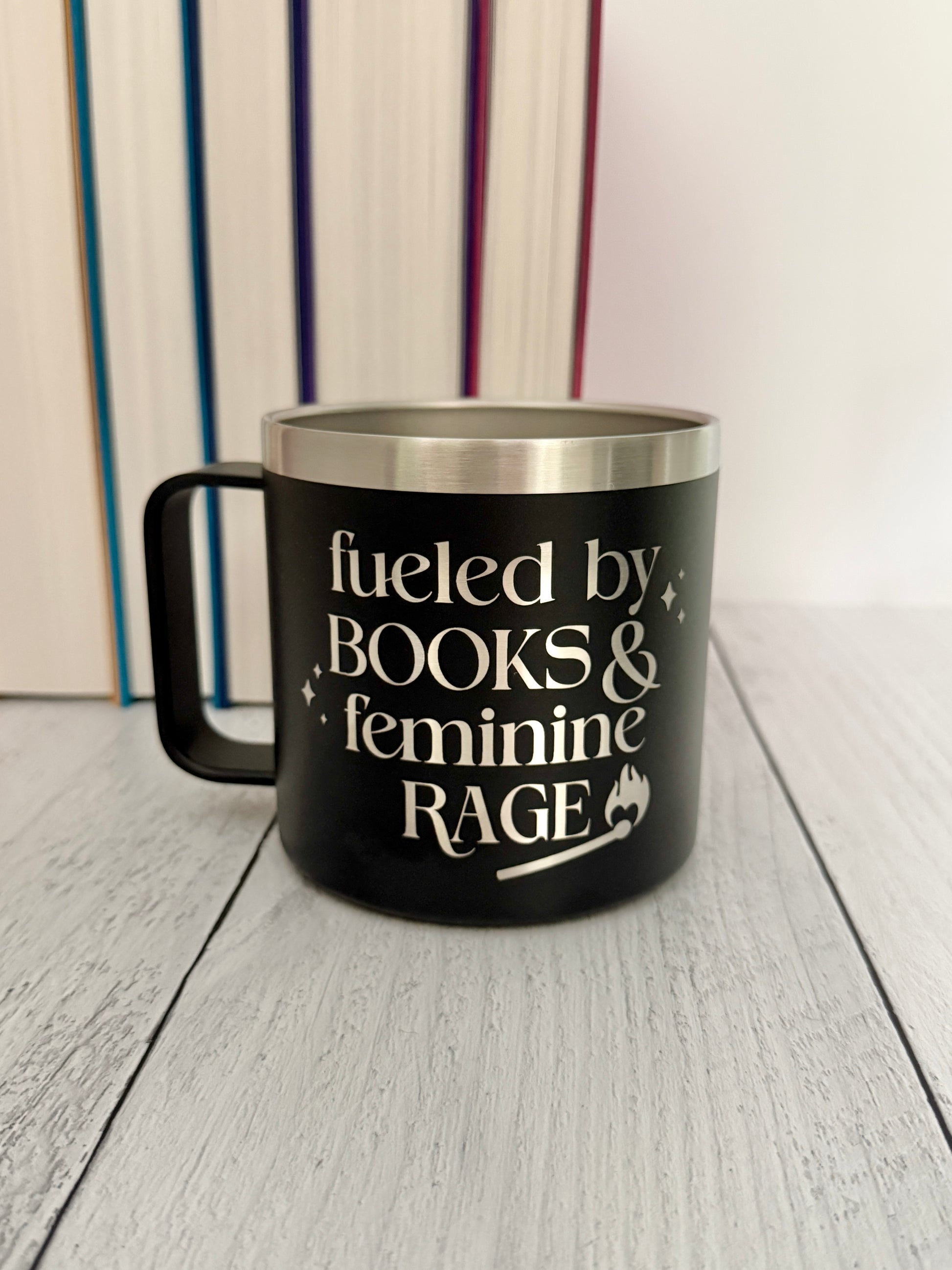 A black insulated travel mug with a stainless steel rim, featuring the engraved text 'fueled by BOOKS & feminine RAGE.' The mug is placed on a light gray wooden surface with a blurred background of books.