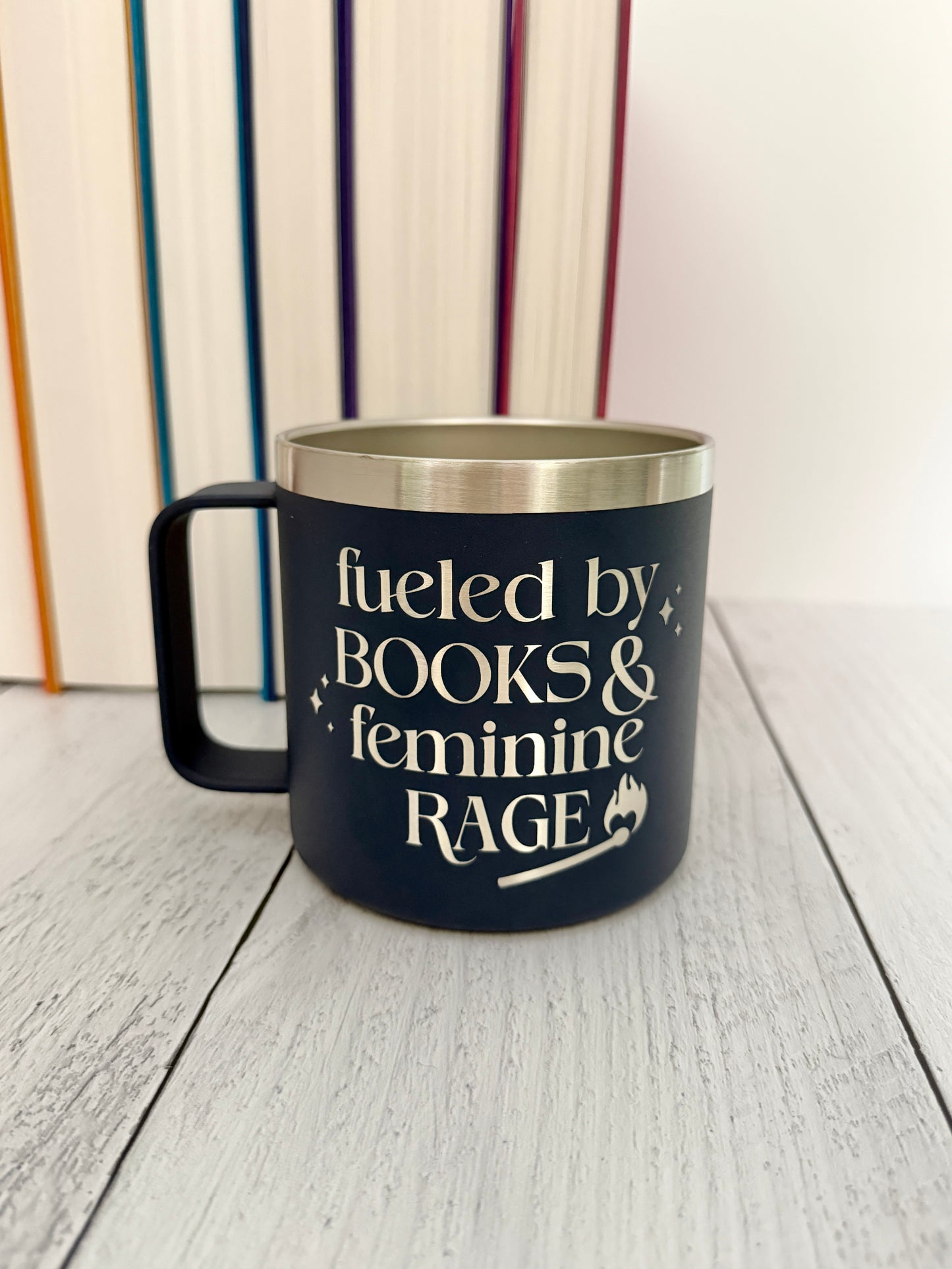 A navy blue insulated mug with a stainless steel rim, featuring the engraved text 'fueled by BOOKS & feminine RAGE.' The mug is placed on a light gray wooden surface with a blurred background of colorful books.