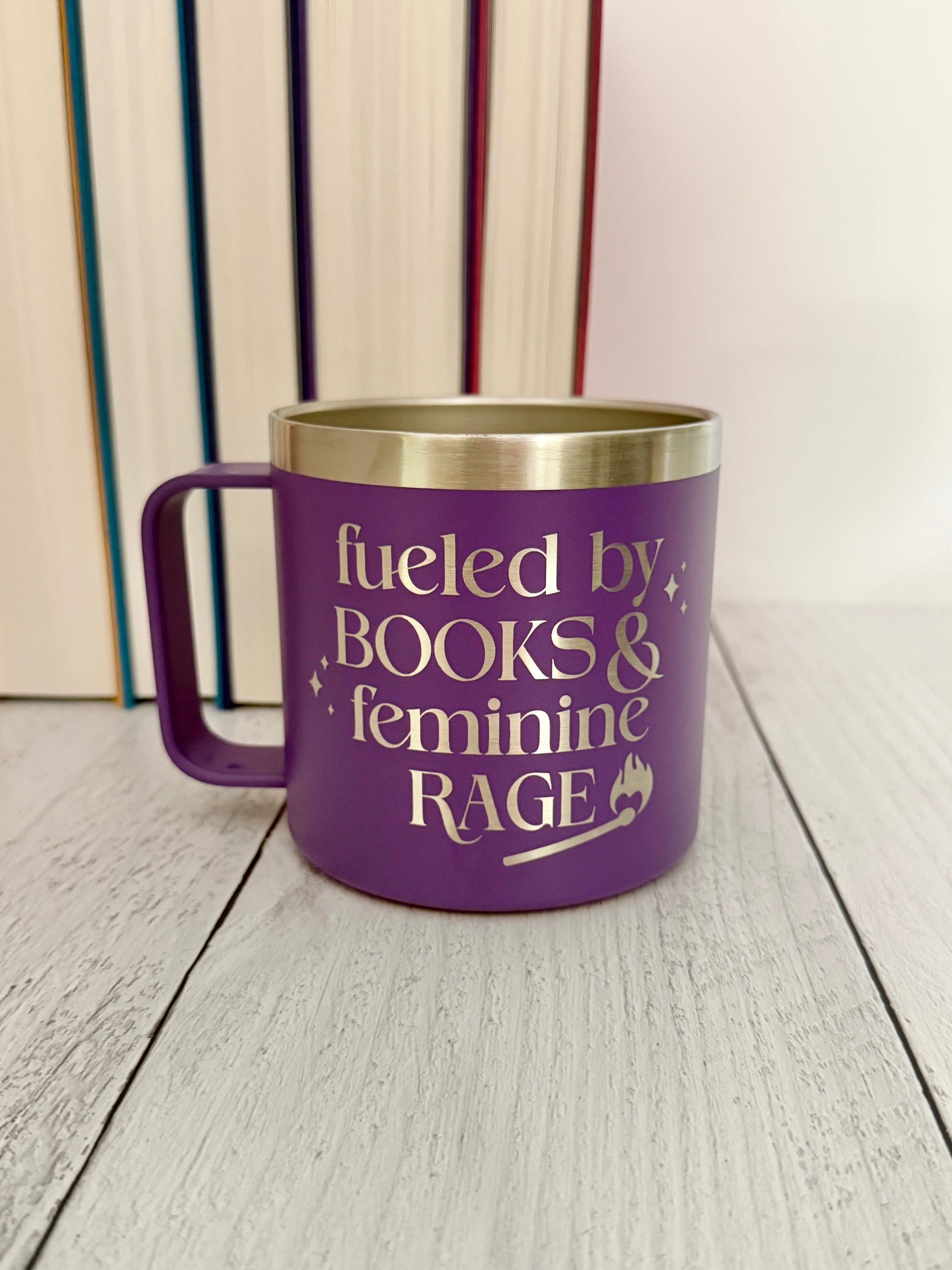 A purple insulated mug with a stainless steel rim and handle, featuring the engraved text 'fueled by BOOKS & feminine RAGE,' set on a light wood surface with books in the background.