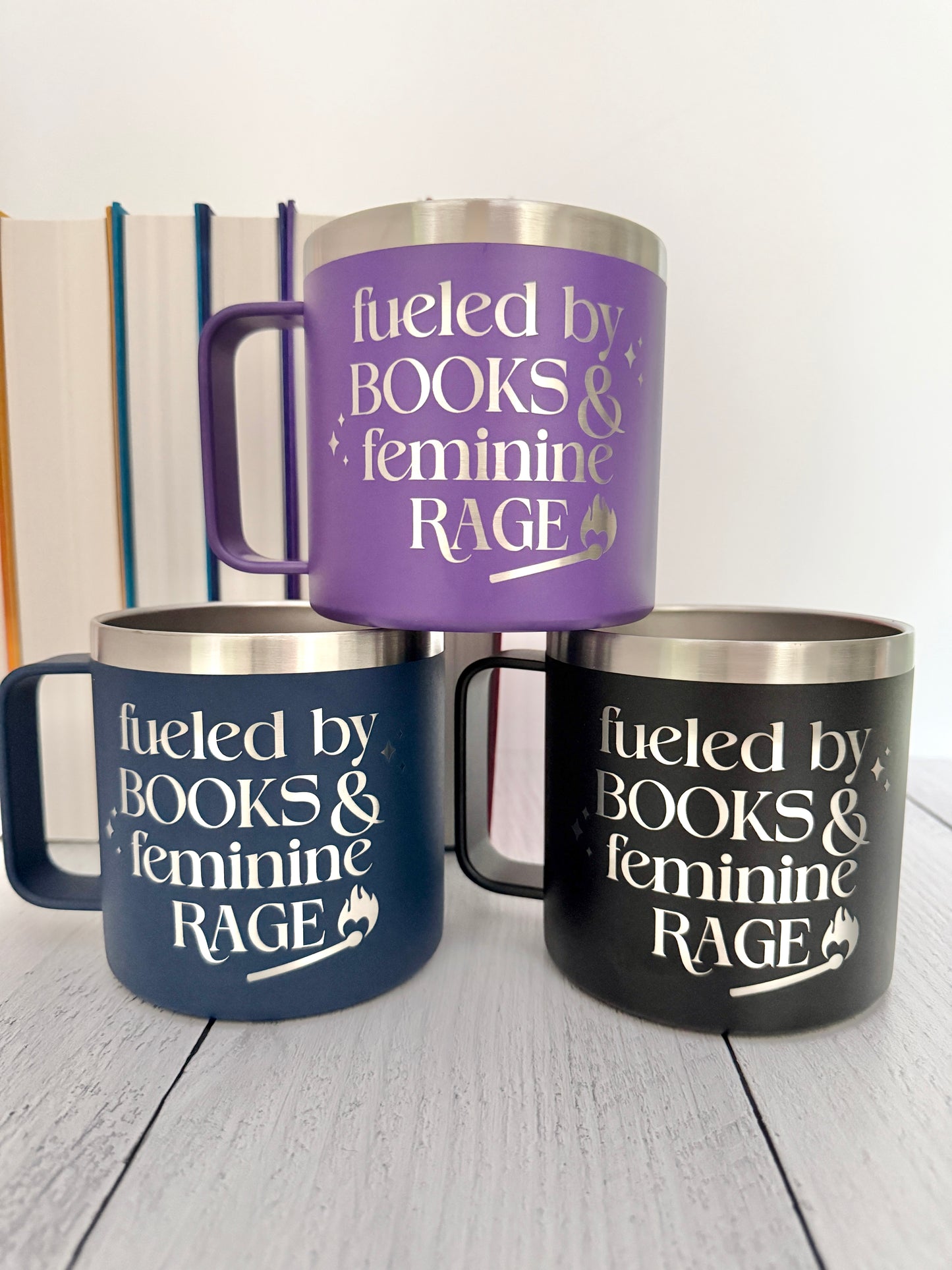 Three insulated travel mugs with stainless steel rims and handles, stacked in a pyramid arrangement on a light wood surface. The top mug is purple, the bottom left is navy blue, and the bottom right is black. They are engraved with the text 'fueled by books and feminine rage.'