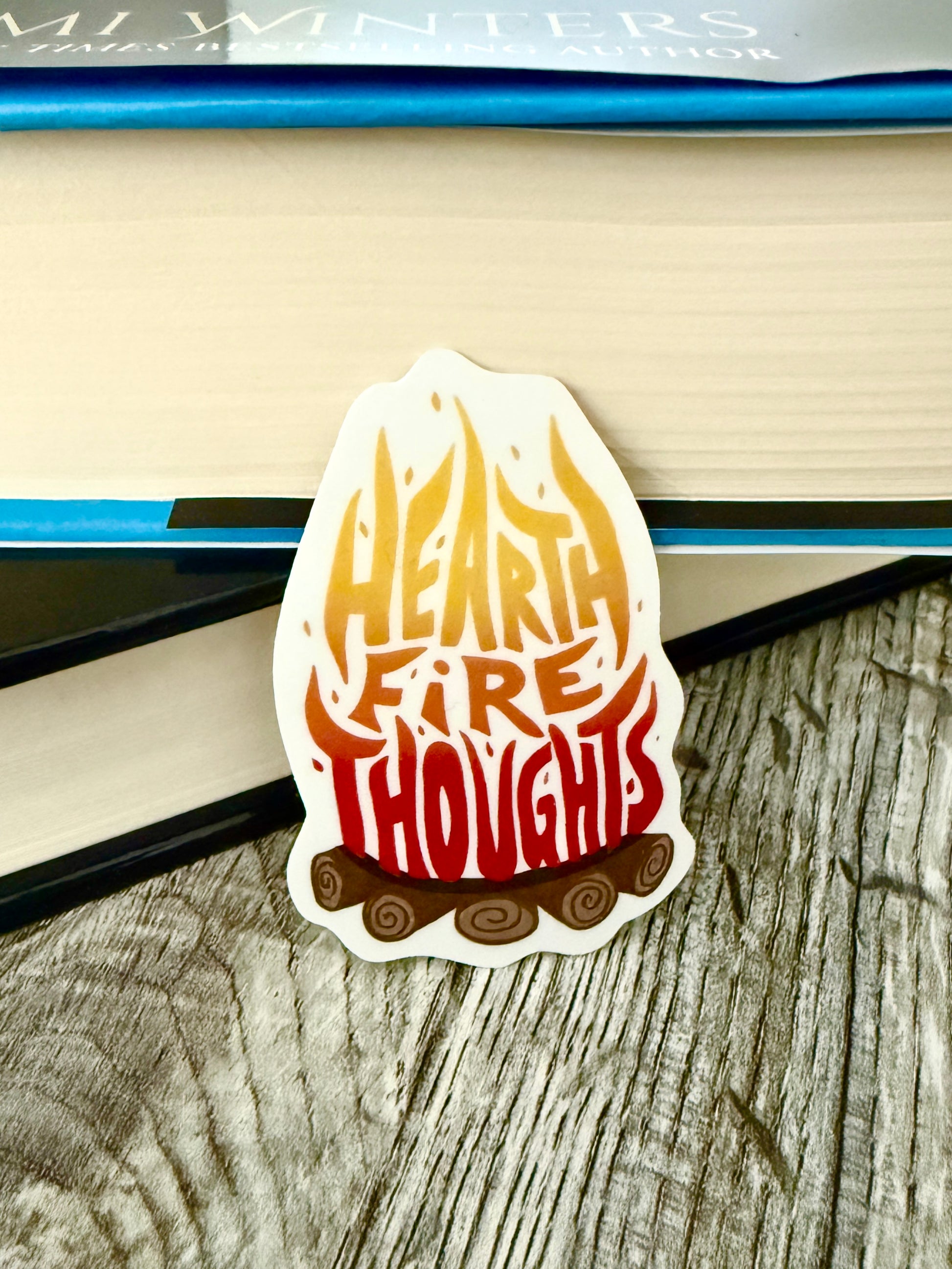 Sticker with 'Heart Fire Thoughts' design, inspired by Road of Bones by Demi Winters, leaning against a book