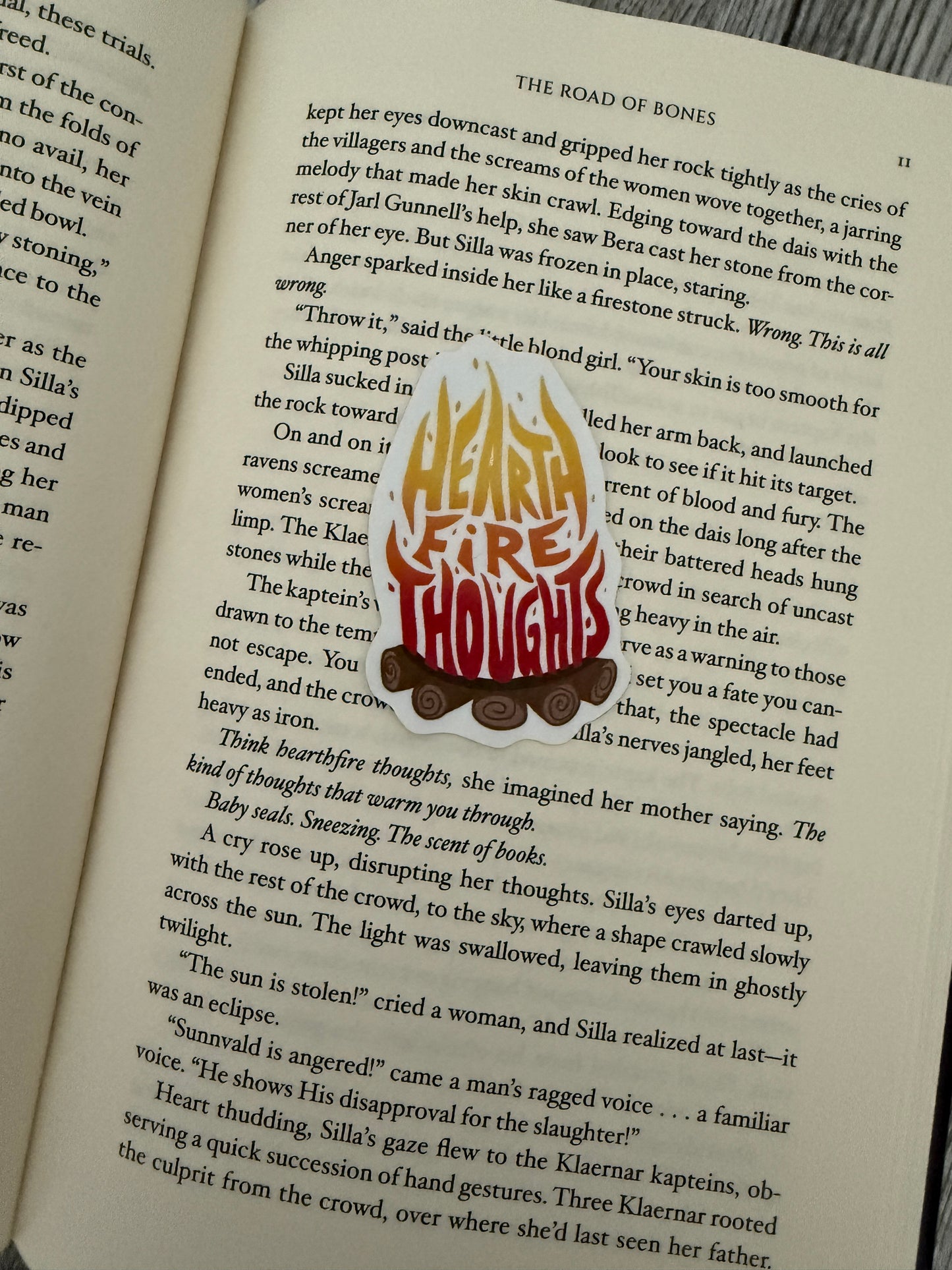 A sticker with "hearth fire thoughts" written in a camp fire, inspired by The Road of Bones by Demi Winter, laying on an open book.