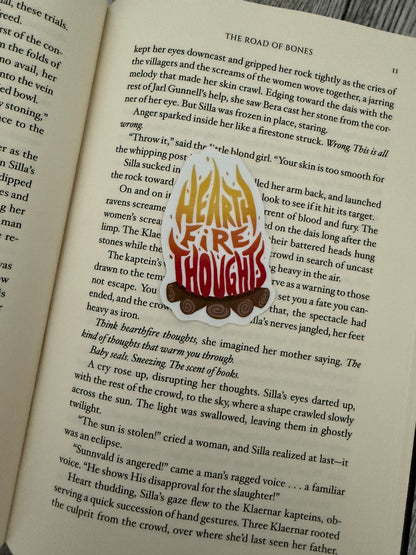 A sticker with "hearth fire thoughts" written in a camp fire, inspired by The Road of Bones by Demi Winter, laying on an open book.