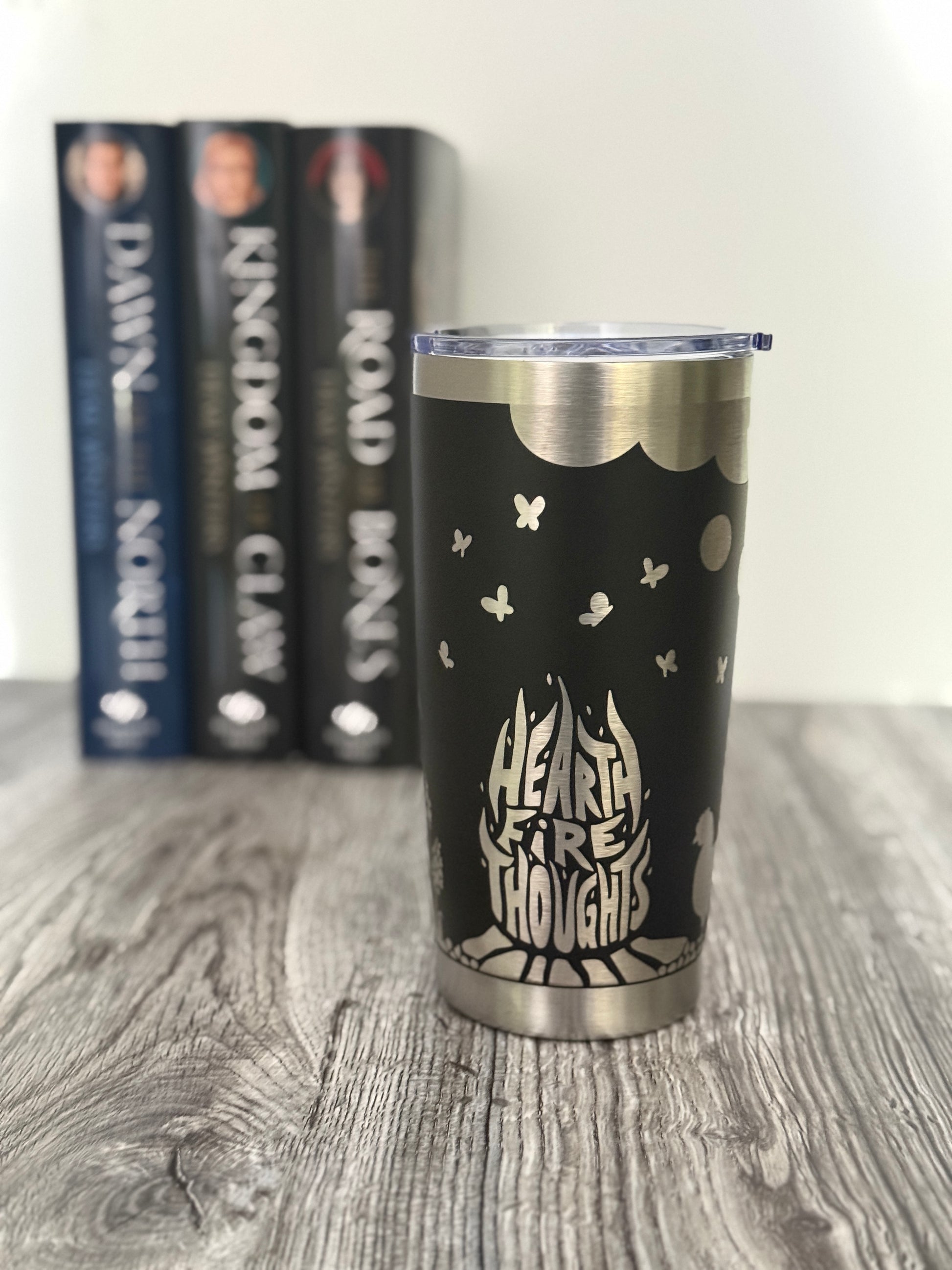 Black tumbler with 'Hearthfire Thoughts' design on a wooden surface with Demi Winters' Ashen series books in the background.
