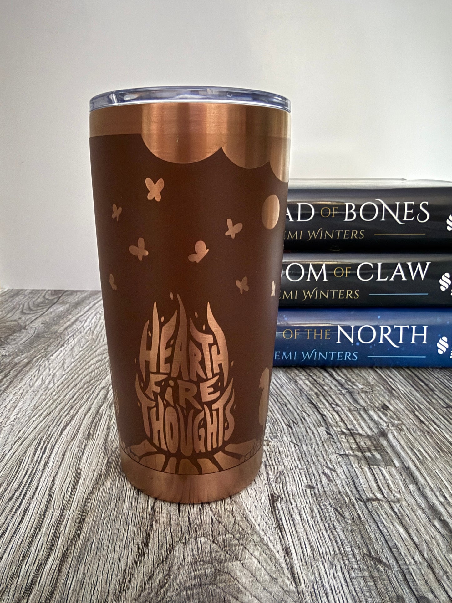 Brown tumbler with 'Heart Fire Thoughts' design on a wooden surface with Demi Winters Road of Bones books in the background.