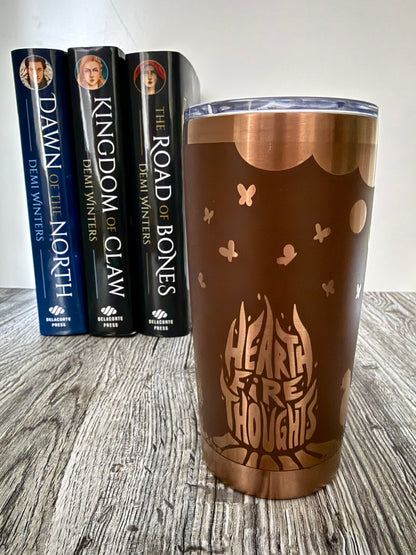 Brown and gold tumbler with 'Hearthfire Thoughts' design in front of Demi Winters' Ashen series books on a wooden surface