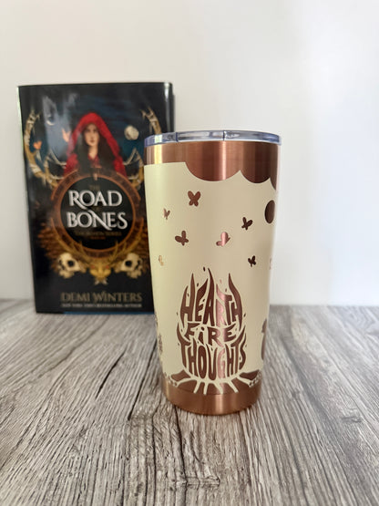 Ivory tumbler with 'Hearthfire Thoughts' design engraved in copper, inspired by the book 'Road of Bones' by Demi Winters in the background.