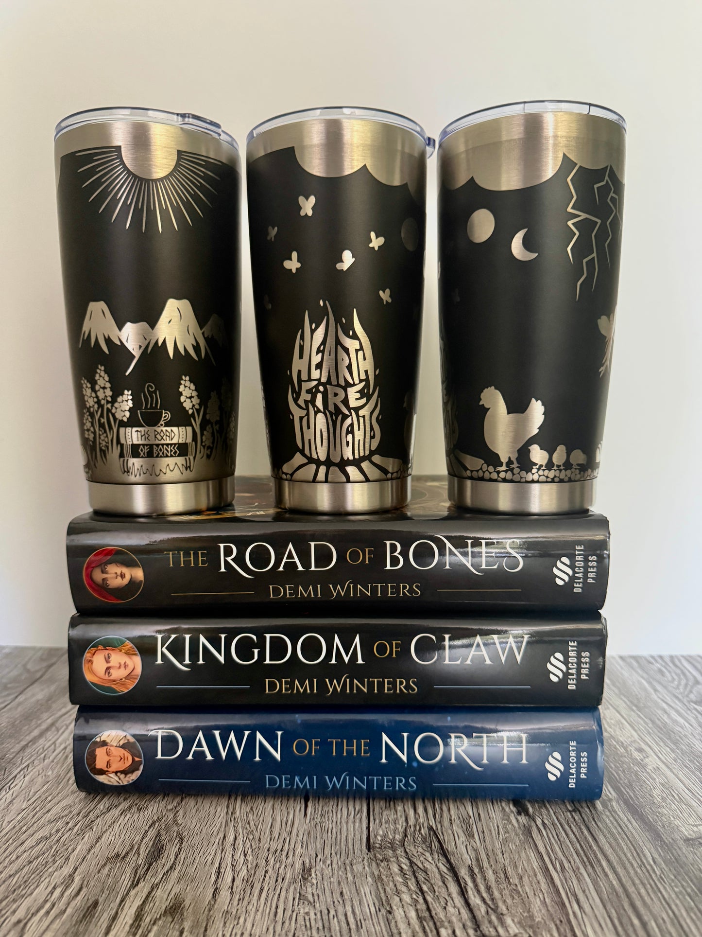 Three black tumblers with engraved 'Hearthfire Thoughts' designs that include chickens, fireweed flowers, snow capped mountains, sunshine, lightning and flita butterflies on top of Demi Winters' Ashen series books: The Road of Bones, Kingdom of Claw, and Dawn of the North.