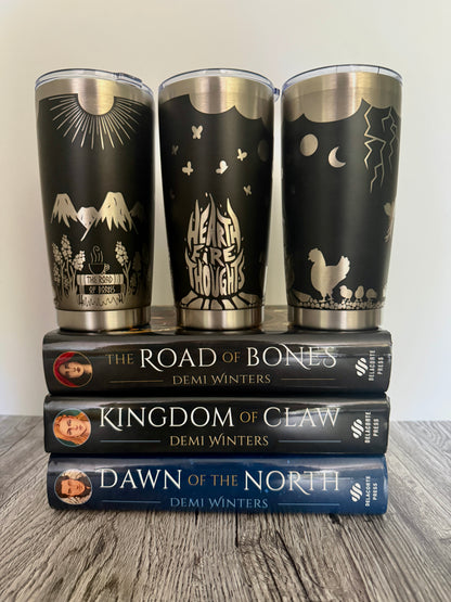 Three black tumblers with engraved 'Hearthfire Thoughts' designs that include chickens, fireweed flowers, snow capped mountains, sunshine, lightning and flita butterflies on top of Demi Winters' Ashen series books: The Road of Bones, Kingdom of Claw, and Dawn of the North.