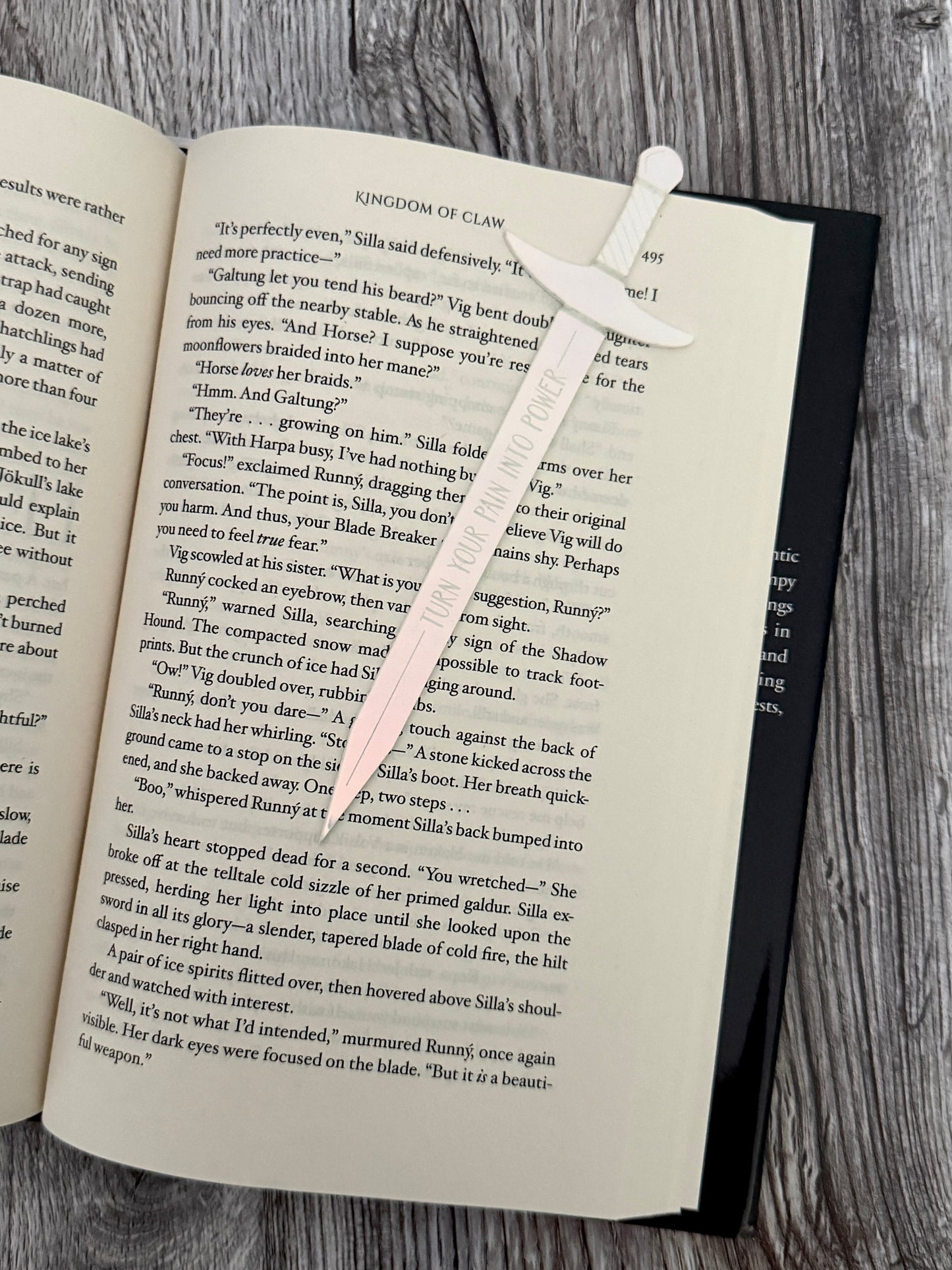 Open book 'Kingdom of Claw' by Demi Winters with a sword shaped bookmark made of iridescent acrylic on a wooden surface