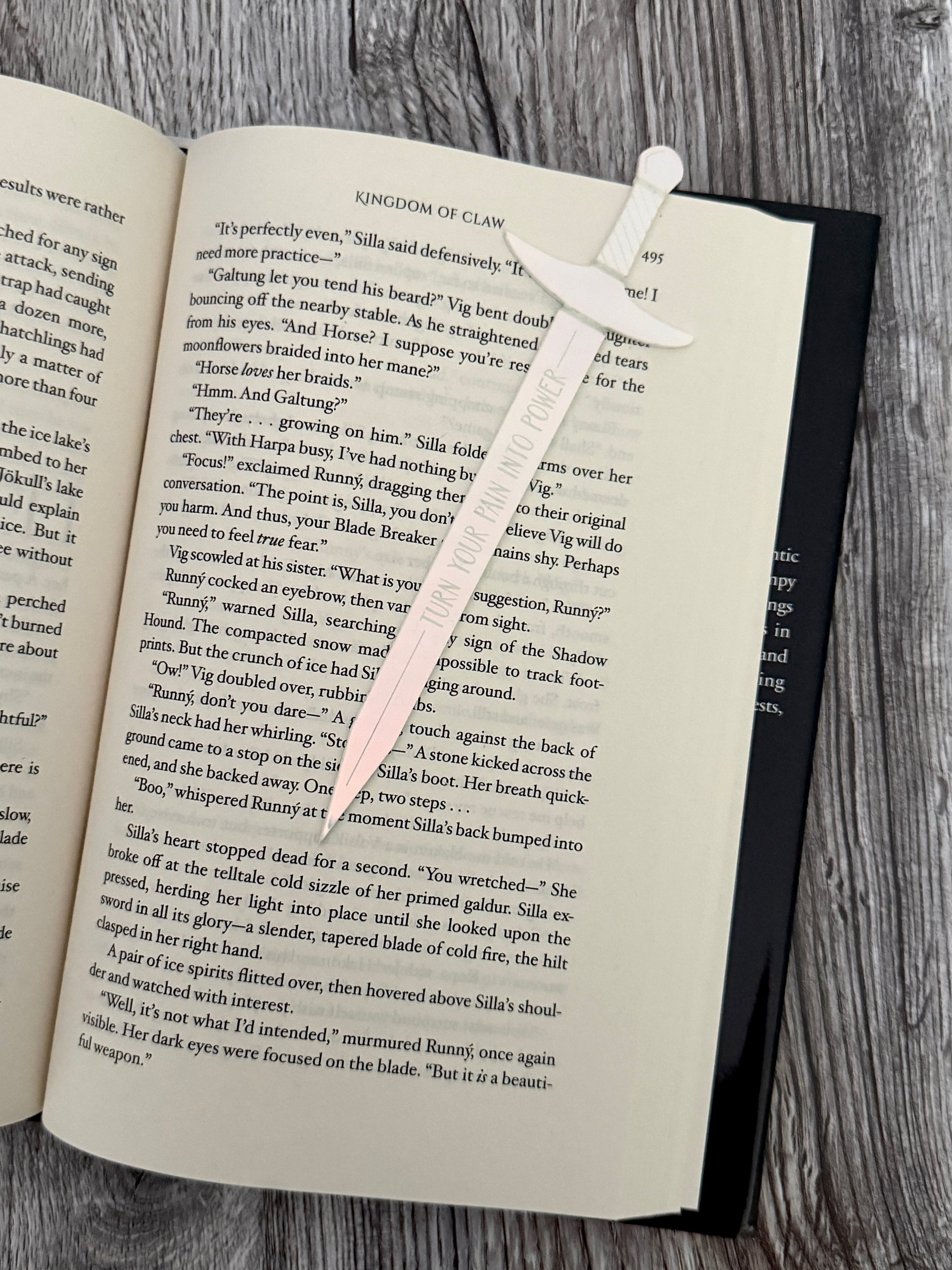 Open book 'Kingdom of Claw' by Demi Winters with a sword shaped bookmark made of iridescent acrylic on a wooden surface