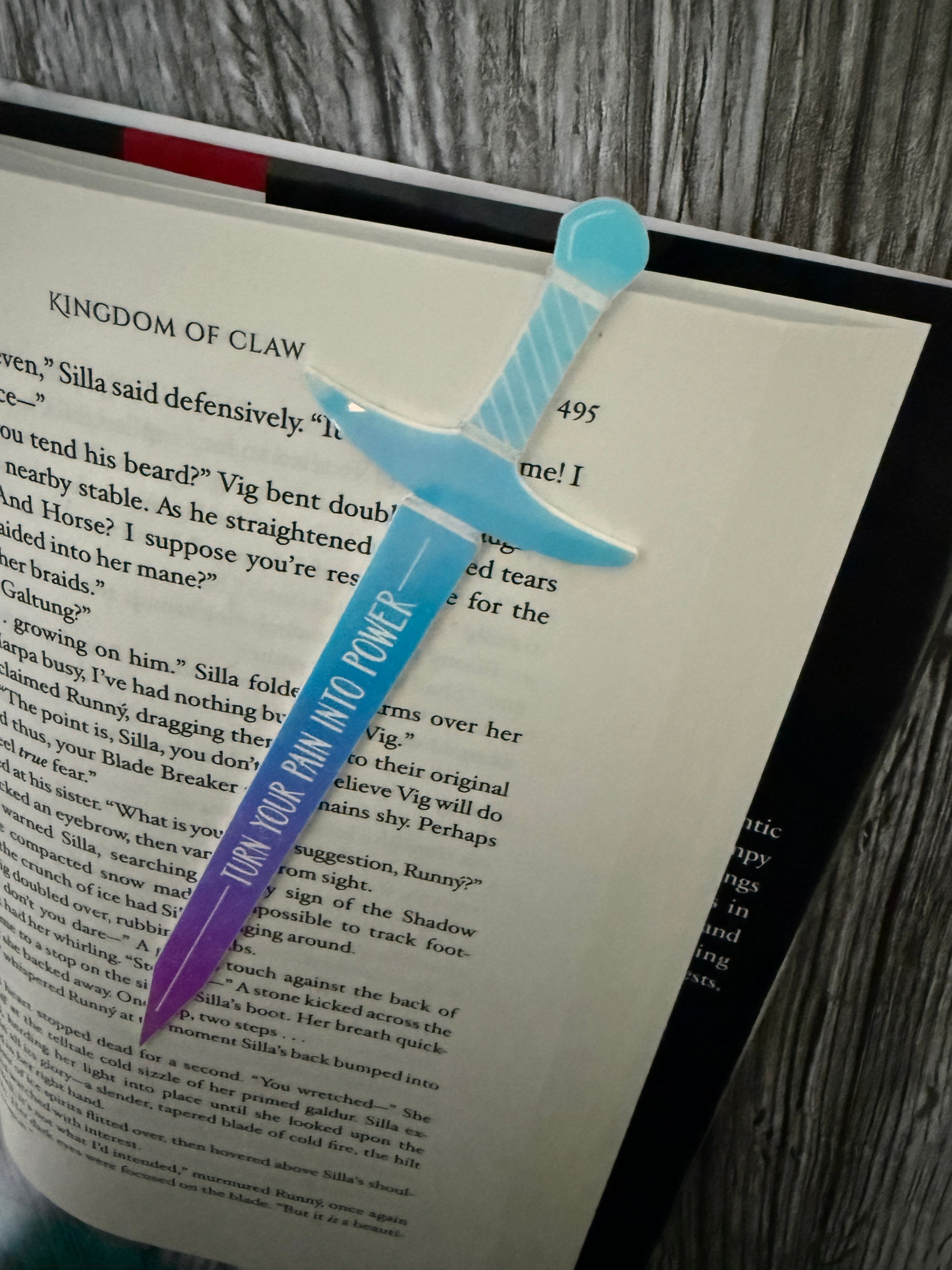 Sword-shaped bookmark with 'turn your pain into Power' text on a 'Kingdom of Claw' book by Demi Winters