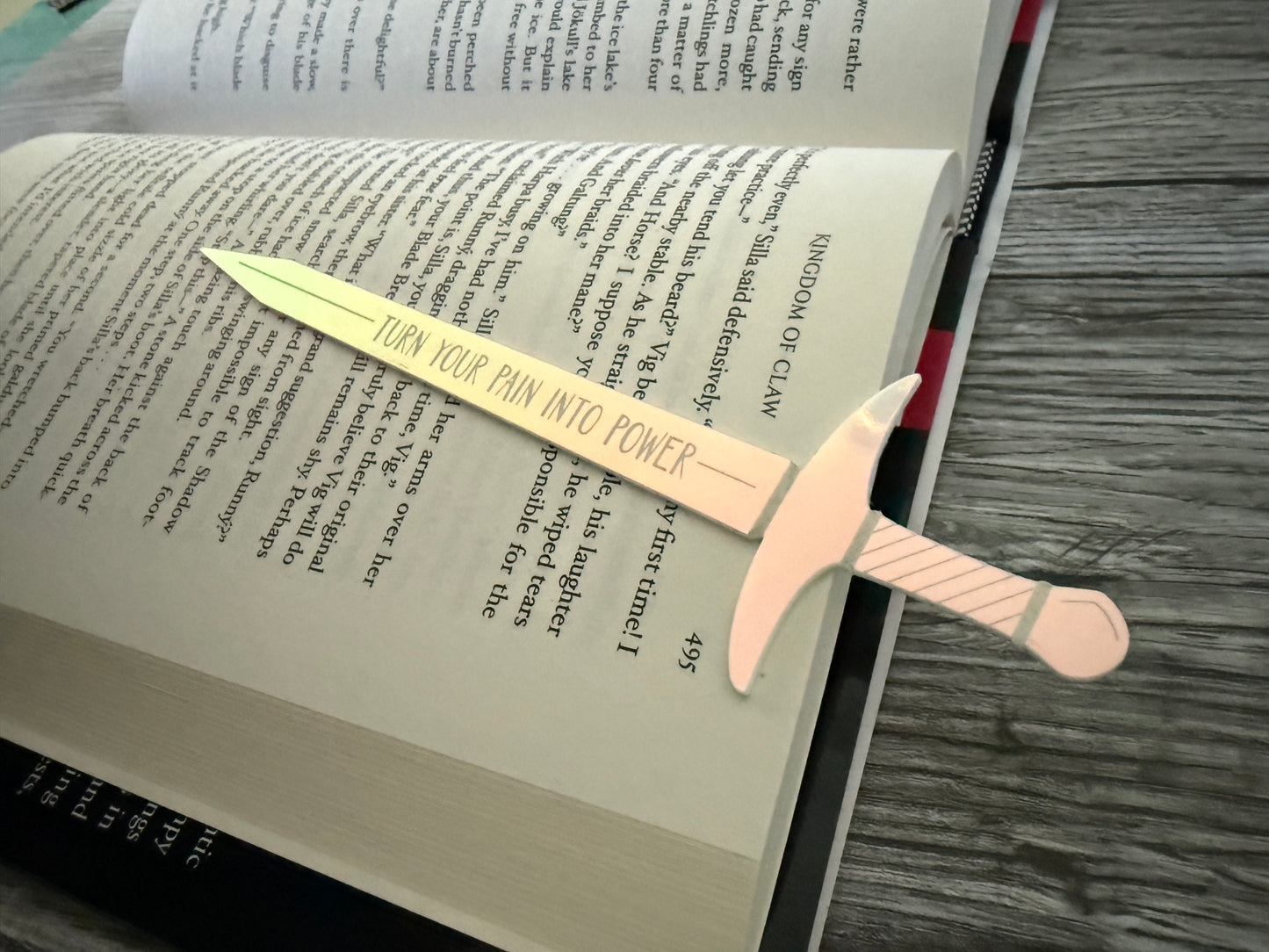Iridescent acrylic sword bookmark engraved with 'turn your pain into power' laying on an open book of 'Kingdom of Claw' by Demi Winters