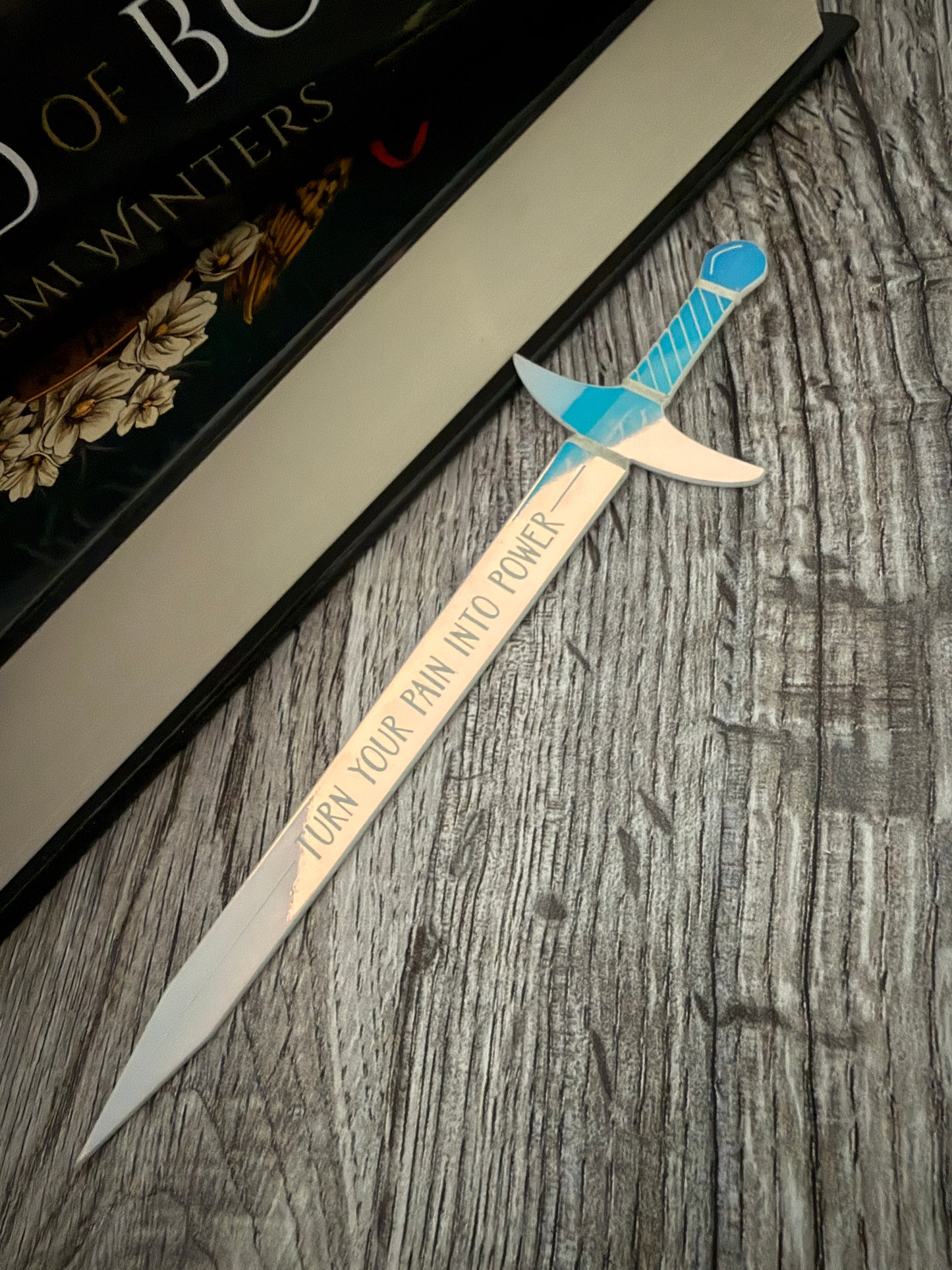 Iridescent sword bookmark engraved with 'turn your pain into power' on a wooden surface, next to 'The Road of Bones' book by Demi Winters