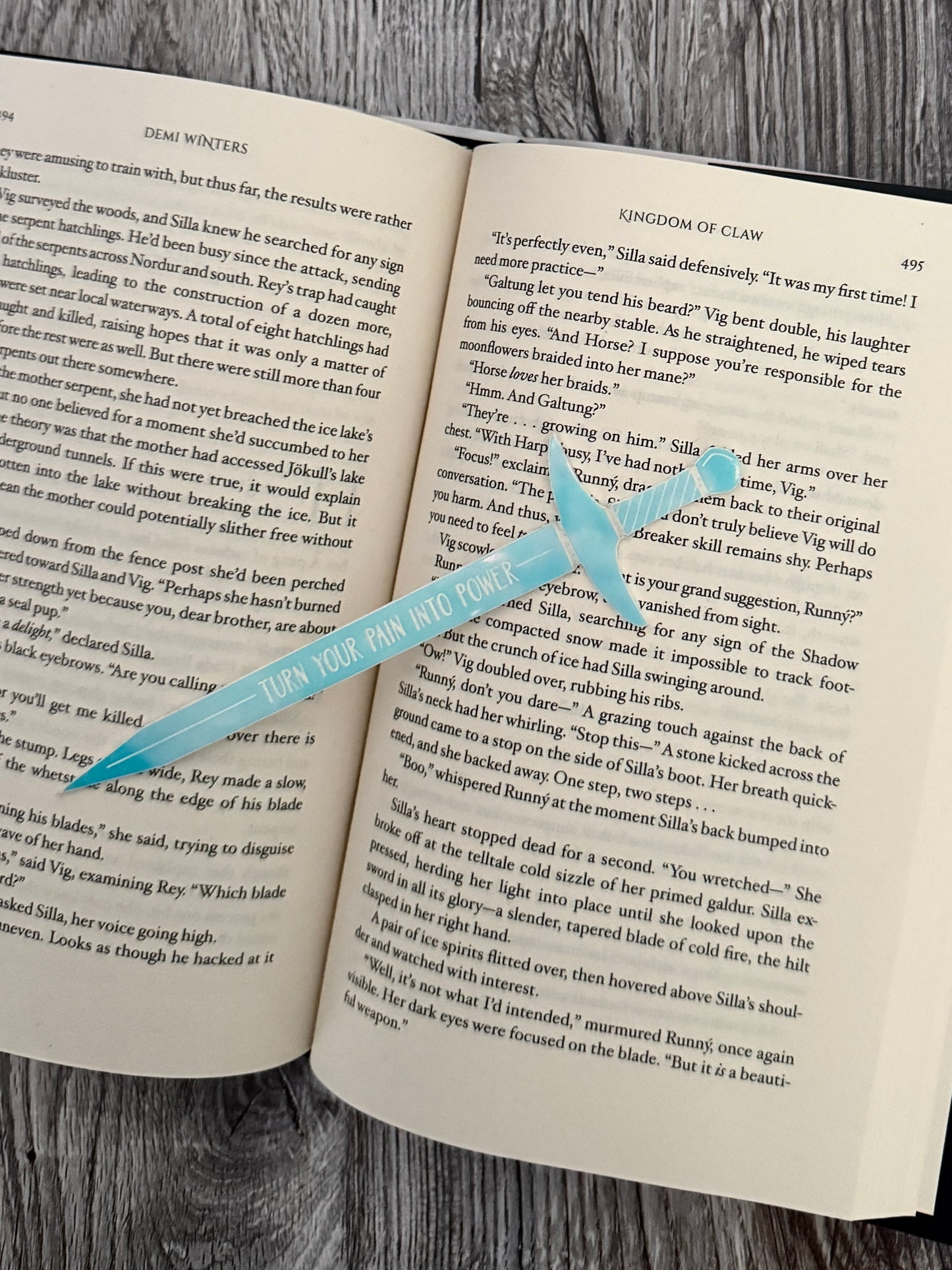 Iridescent sword bookmark engraved with 'turn your pain into power' on an open book of 'Kingdom of Claw' by Demi Winters