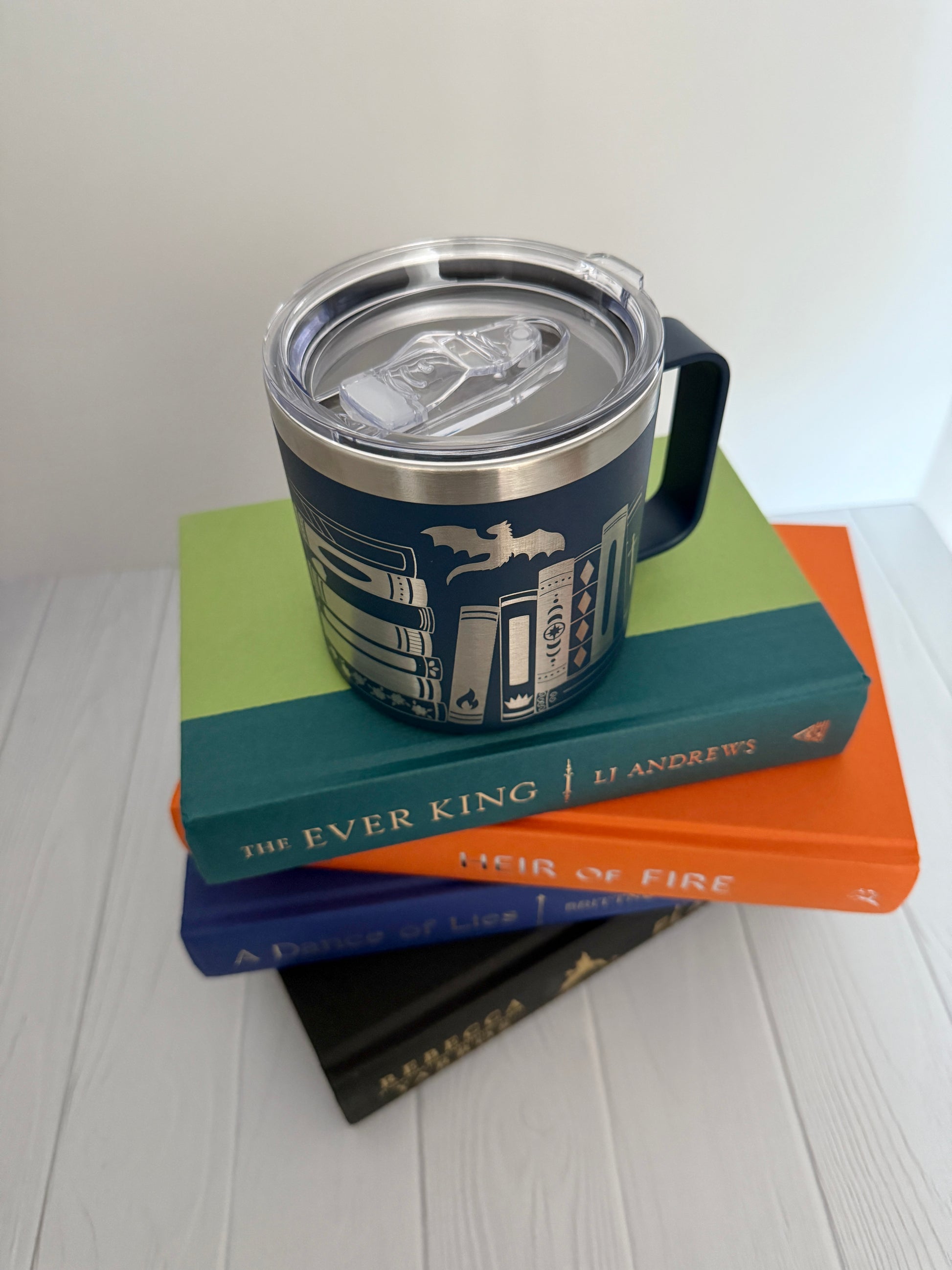 Blue mug with engraved books and dragon featuring a clear lid on top of stack of books on a white surface