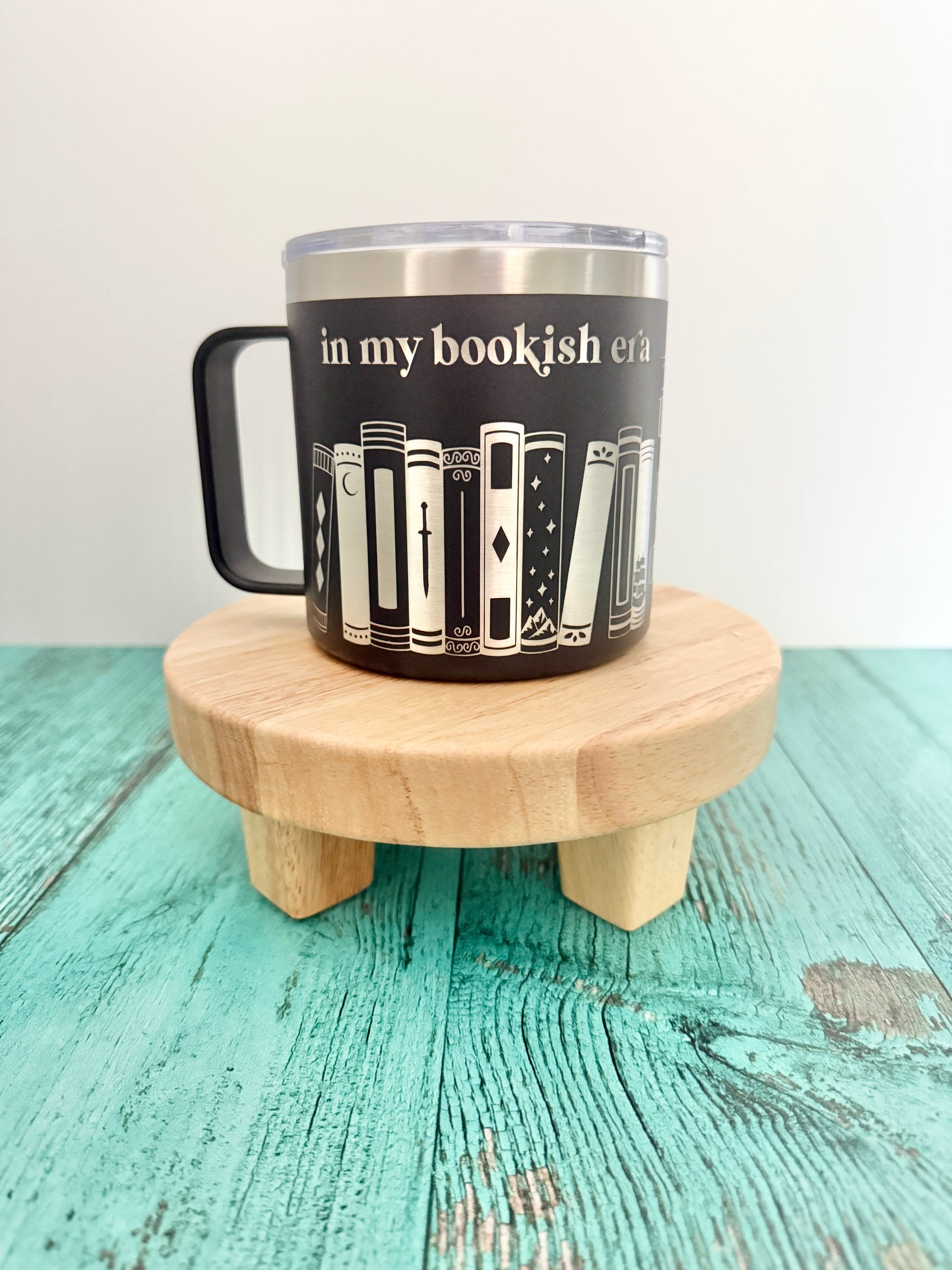 Black mug with book design engraved 'in my bookish era' on a wooden coaster.