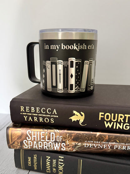 black stainless steel mug engraved with 'in my bookish era' above books on a shelf, set atop a stack of black and gold books.