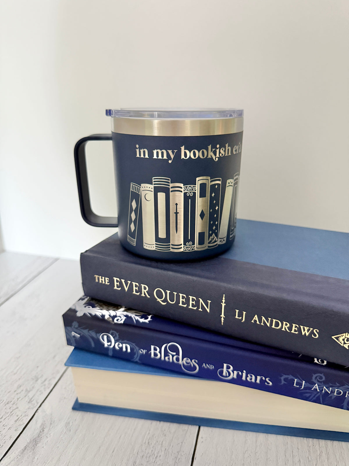 Navy stainless steel mug with 'in my bookish era' and books engraved around it, set on top of a stack of blue books.