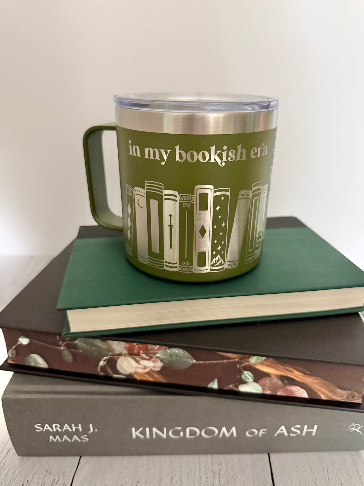 Green stainless steel mug with 'in my bookish era' and books engraved around it, set on top of a stack of green , black and gray books.
