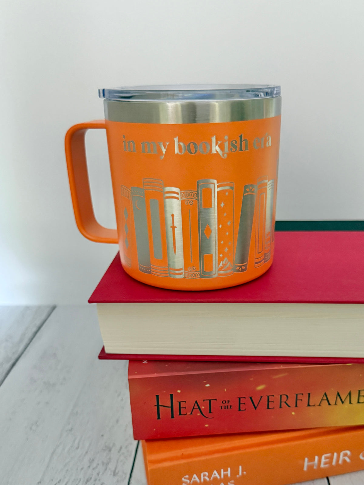 Orange stainless steel mug with 'in my bookish era' and books engraved around it, set on top of a stack of orange and red books.